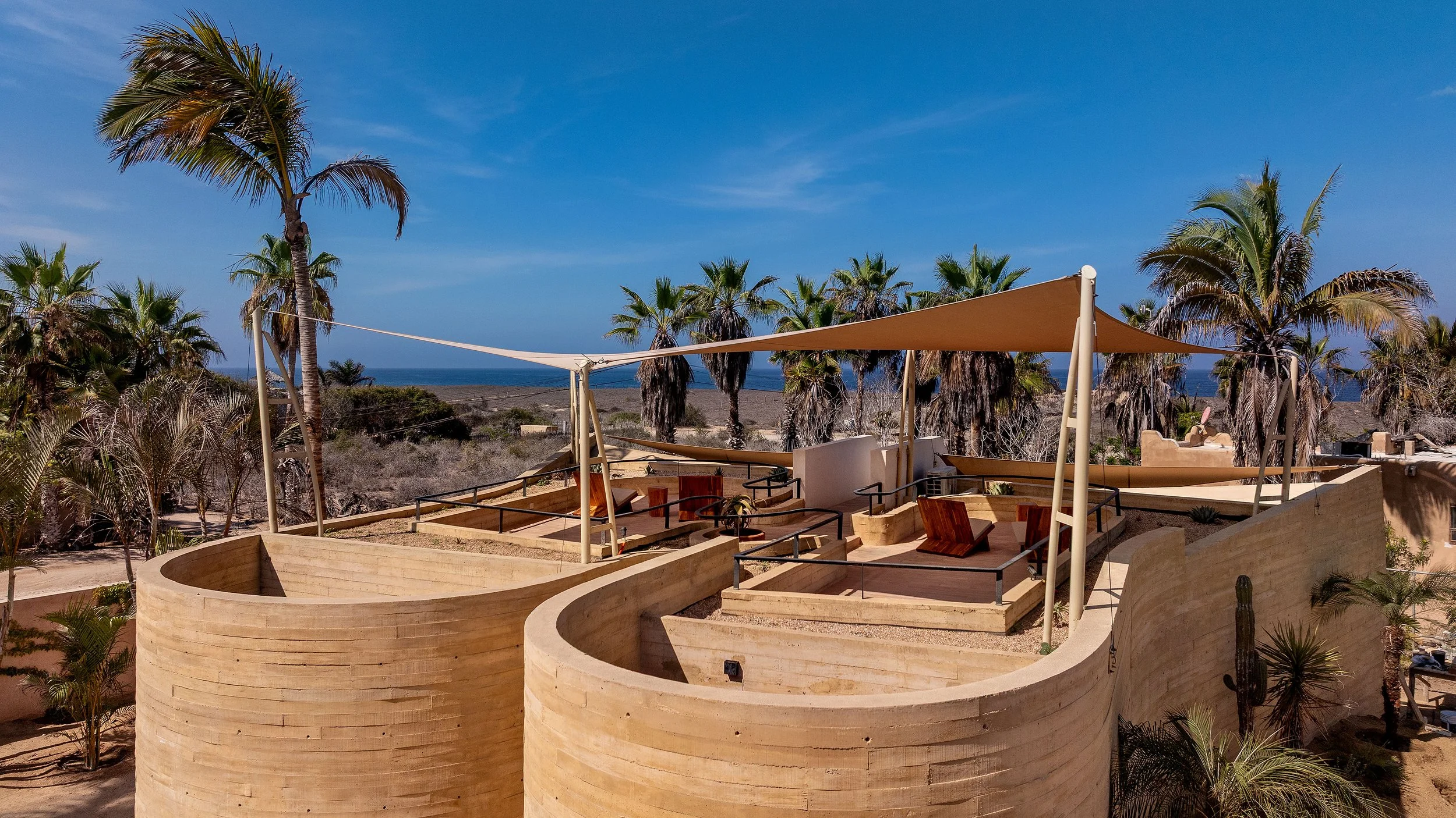 Outdoor rooftop lounge area with wooden chairs and shaded sails, surrounded by palm trees, overlooking the ocean under a blue sky.