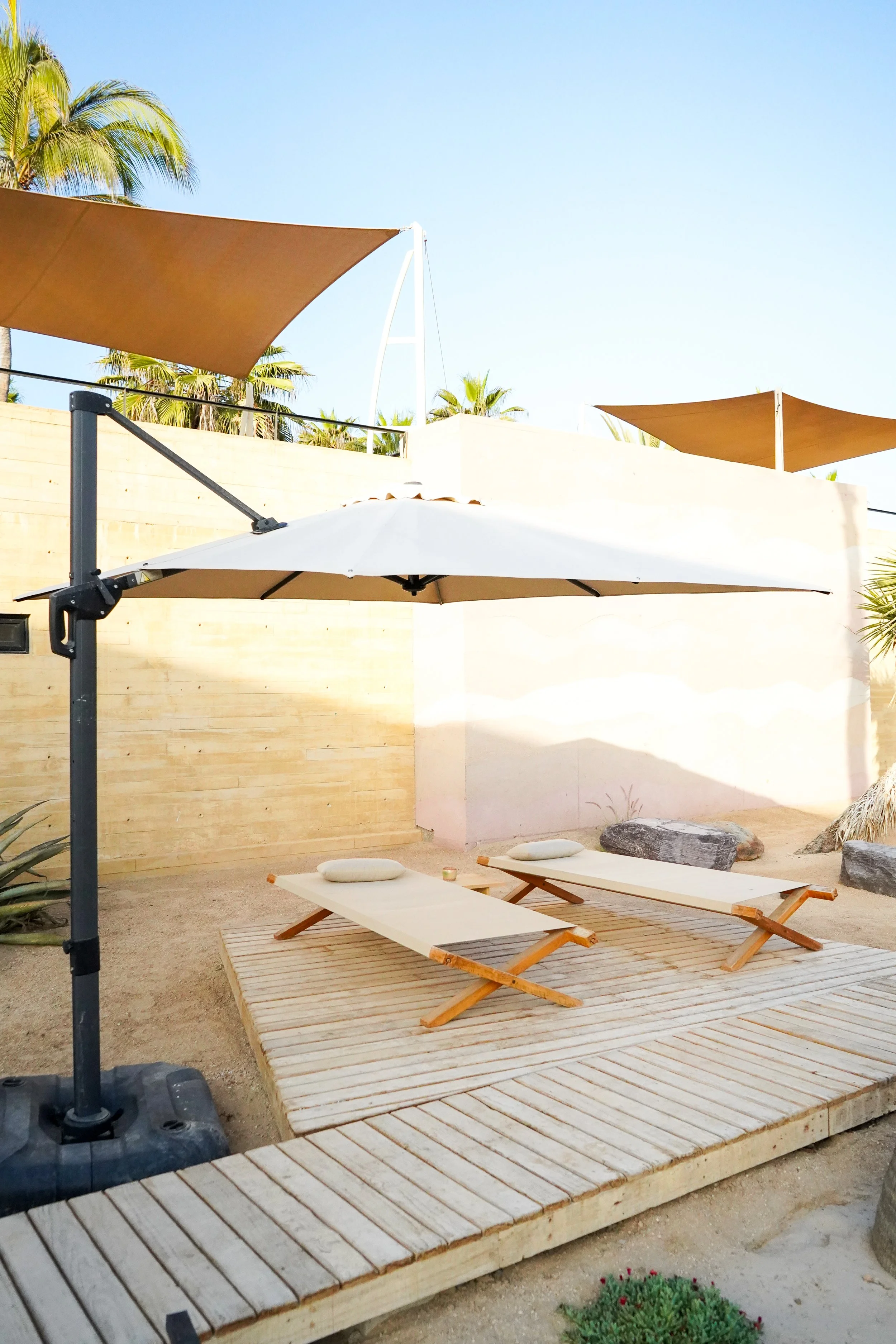 Two lounge chairs with cushions beneath a large white umbrella on a wooden deck, a shaded outdoor area with beige and brown canopies, surrounded by desert landscaping, rocks, and a beige wall with palm trees in the background.