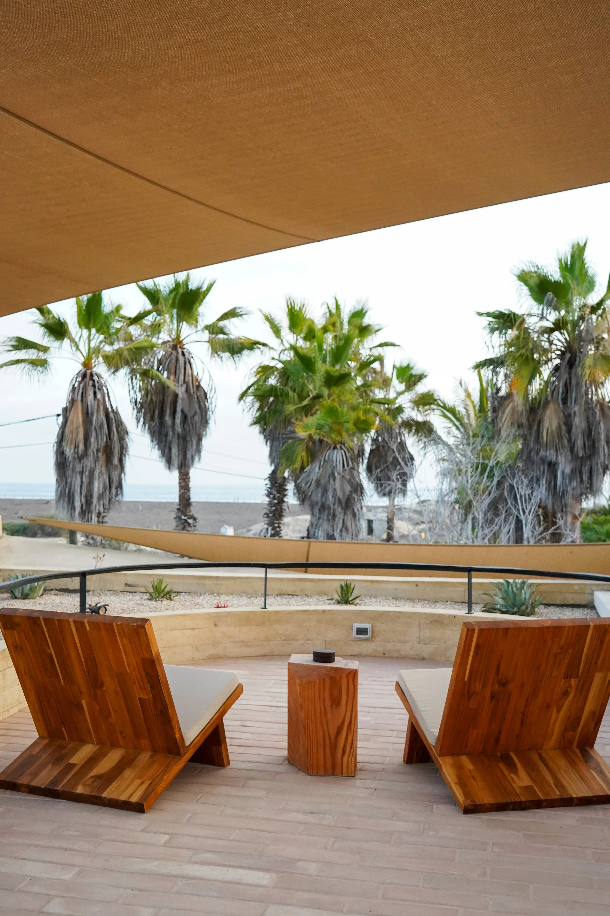 Outdoor patio with two wooden chairs facing a view of palm trees and a sandy beach, under a beige overhang.