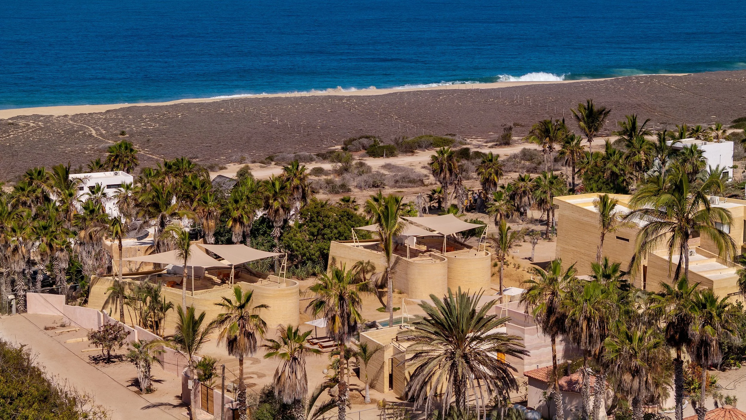 Aerial view of a beachside resort with modern buildings surrounded by palm trees, sandy pathways, and a shoreline with waves in the background.