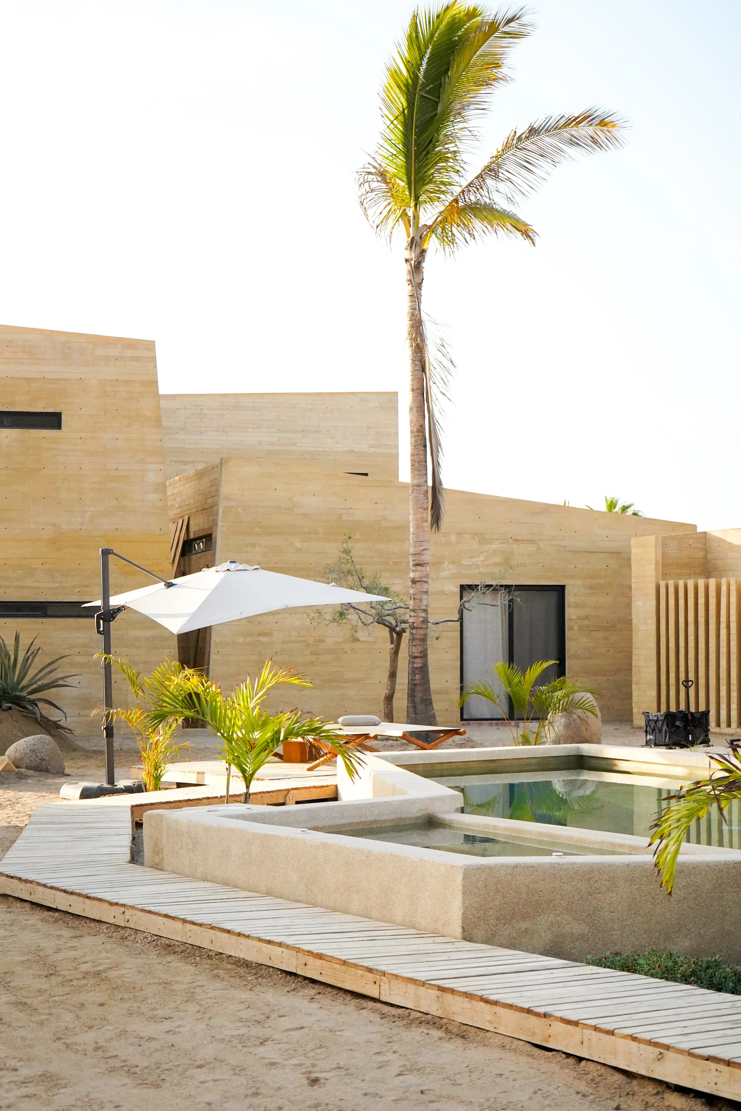 A modern desert home with a swimming pool, palm trees, a white umbrella, and wooden deck, surrounded by desert landscaping.