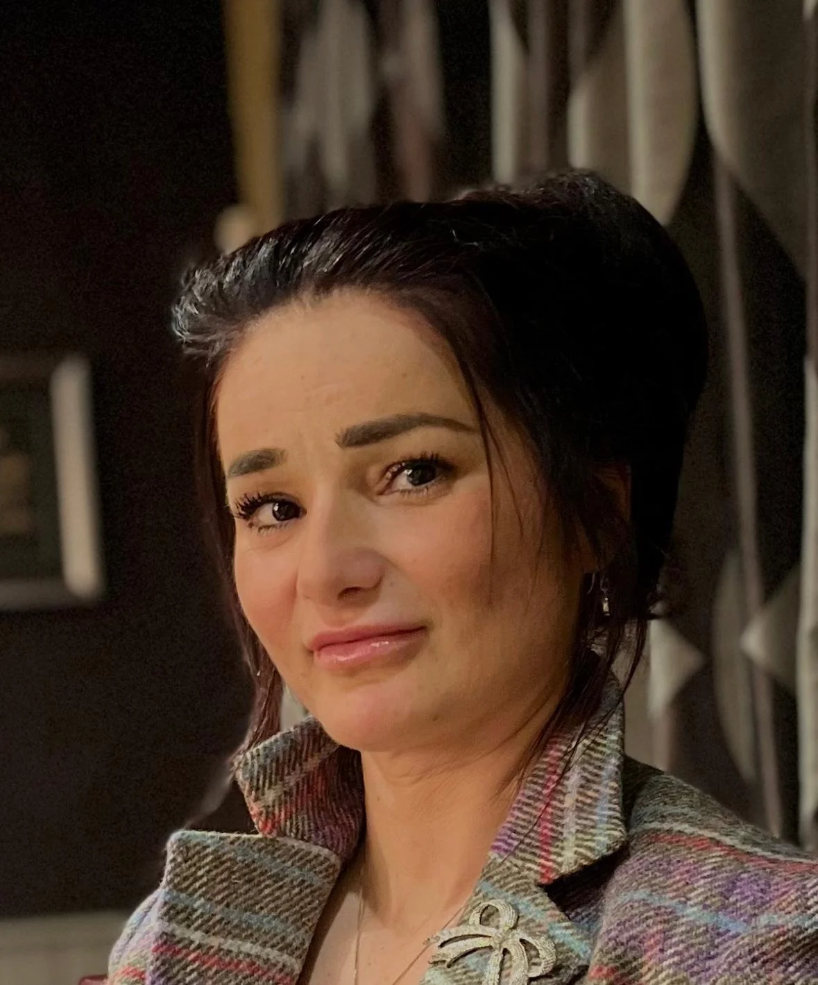 A woman with dark hair styled in an updo, wearing a plaid blazer with a bow accessory, posing indoors with a serious expression.