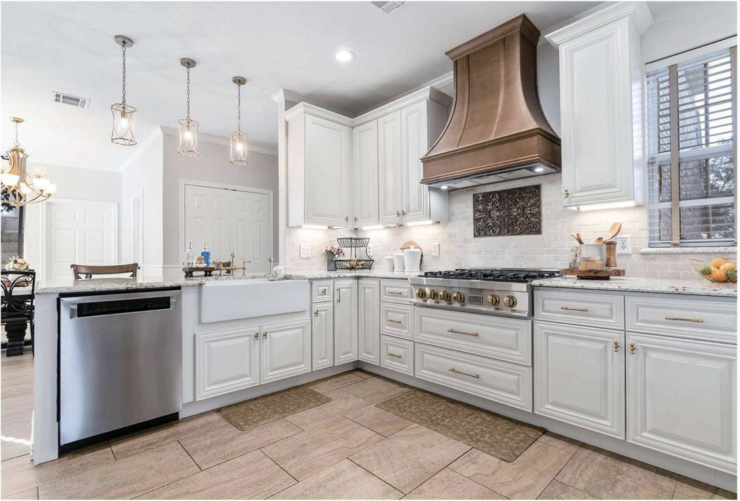 A modern kitchen with white cabinets, a large stove with an ornate range hood, granite countertops, and tiled flooring. There are pendant lights hanging from the ceiling and a window with blinds on the right side.