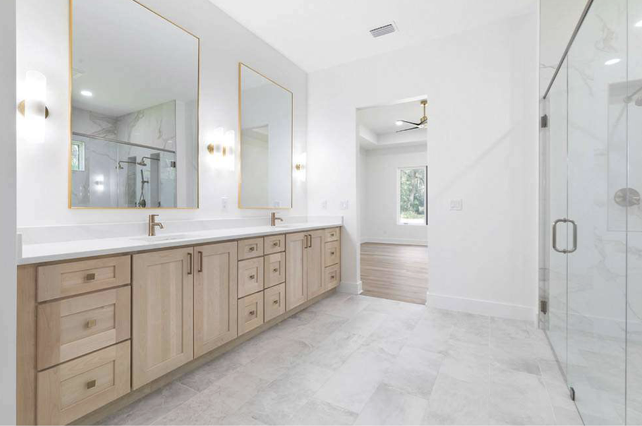 Modern bathroom with double vanity, large mirrors, gold fixtures, glass shower enclosure, and bright white walls.