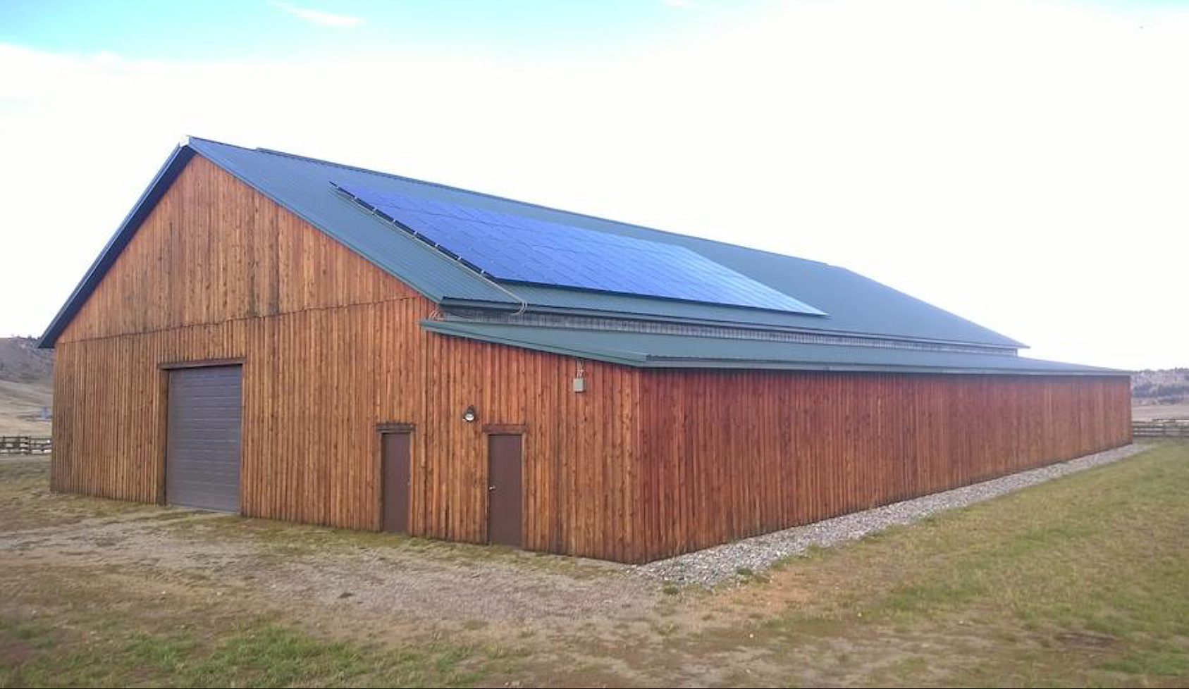 A large wooden barn with a sloped metal roof, equipped with solar panels, situated in a rural landscape with grass and open fields.
