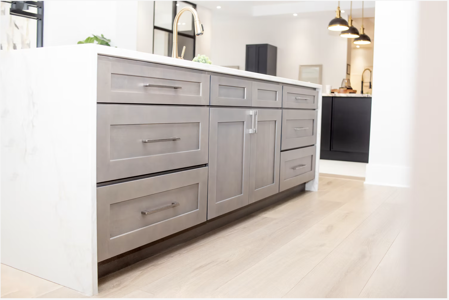 Modern kitchen island with gray cabinetry, white countertop, and built-in drawers and cabinets, with a brass faucet and kitchen decor in the background.