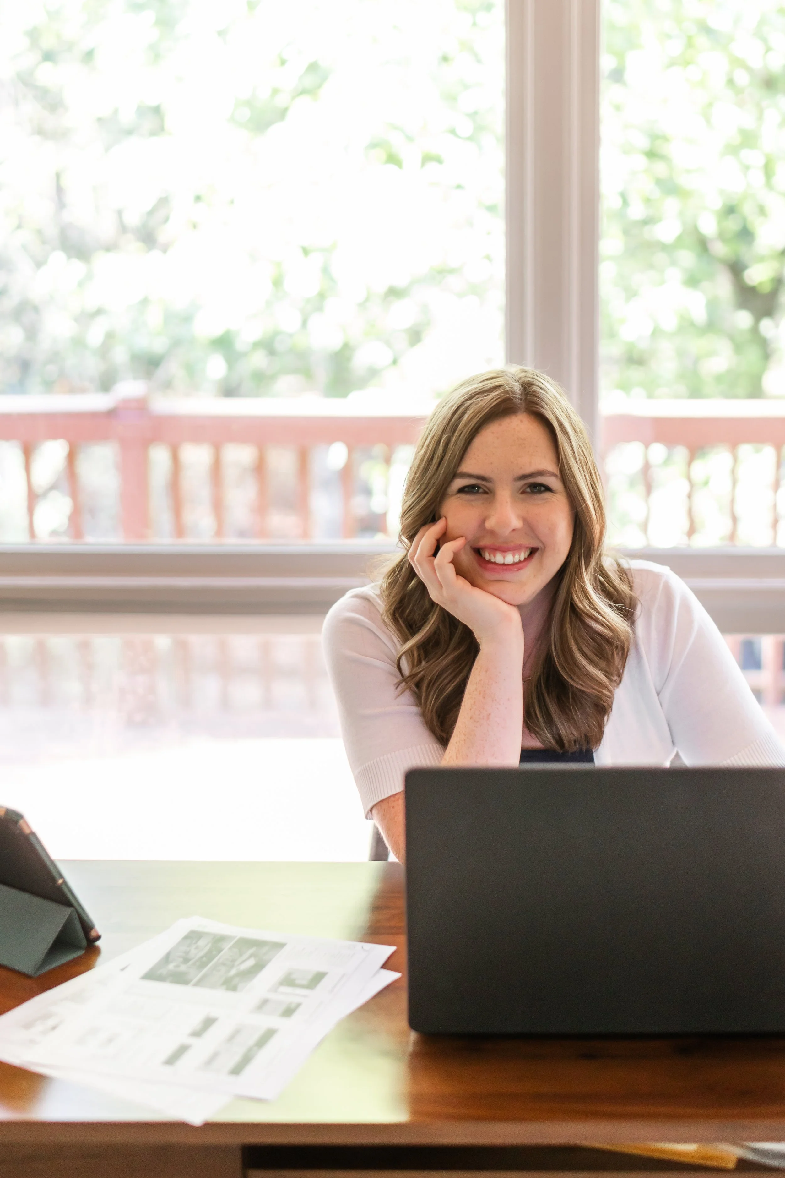 A woman with long brown hair and freckles smiling at the camera, sitting at a work desk with a laptop, papers, and a tablet in front of her, in a room with large windows and greenery outside.