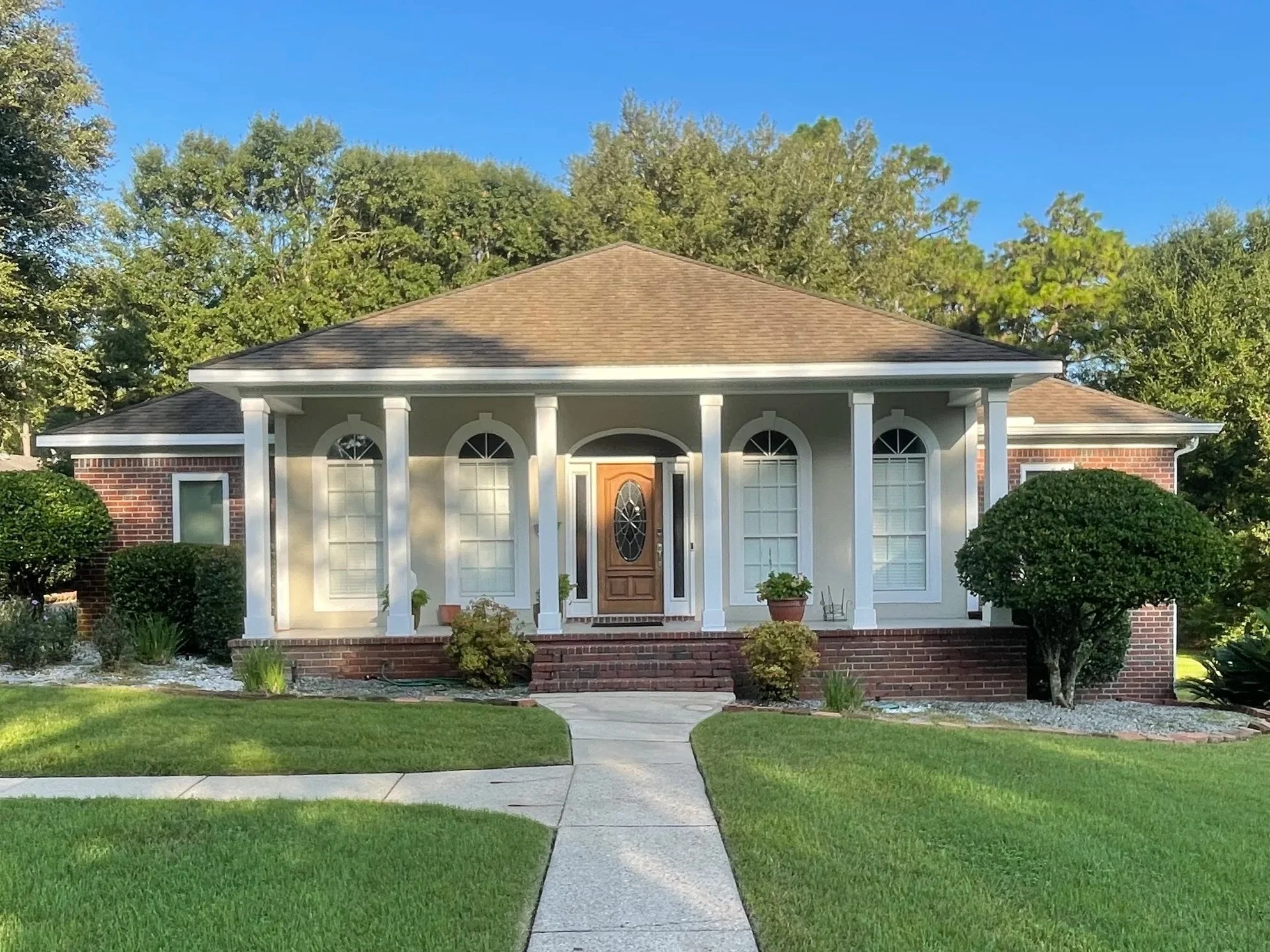 Front view of a house with a brick and white exterior, large arched windows, a wooden front door, a curved walkway, and a well-maintained lawn with bushes and trees in the background.