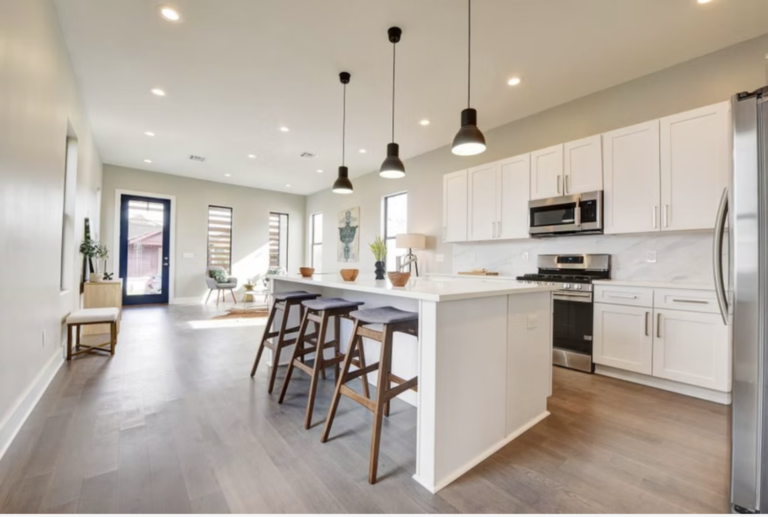 Bright modern kitchen with white cabinets, stainless steel appliances, and a large island with three barstools. Pendant lights hang over the island. A small dining area is visible in the background with large windows and a door leading outside.