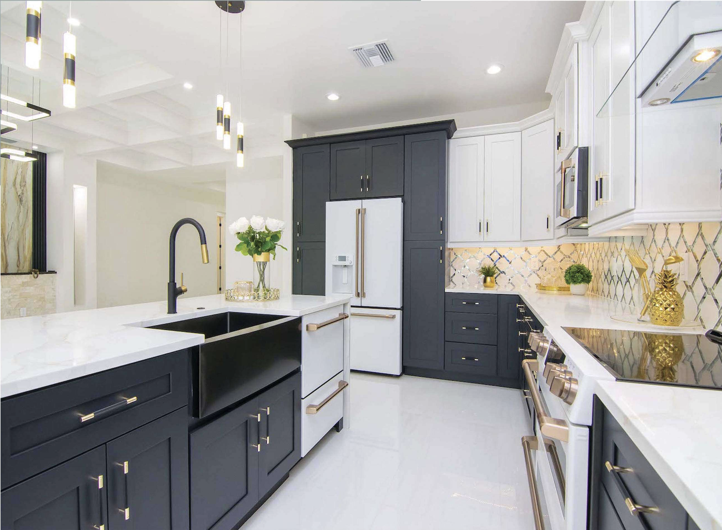 Modern kitchen with navy blue and white cabinets, gold accents, a white marble island with a sink, gold and black pendant lights, and decorative plants.