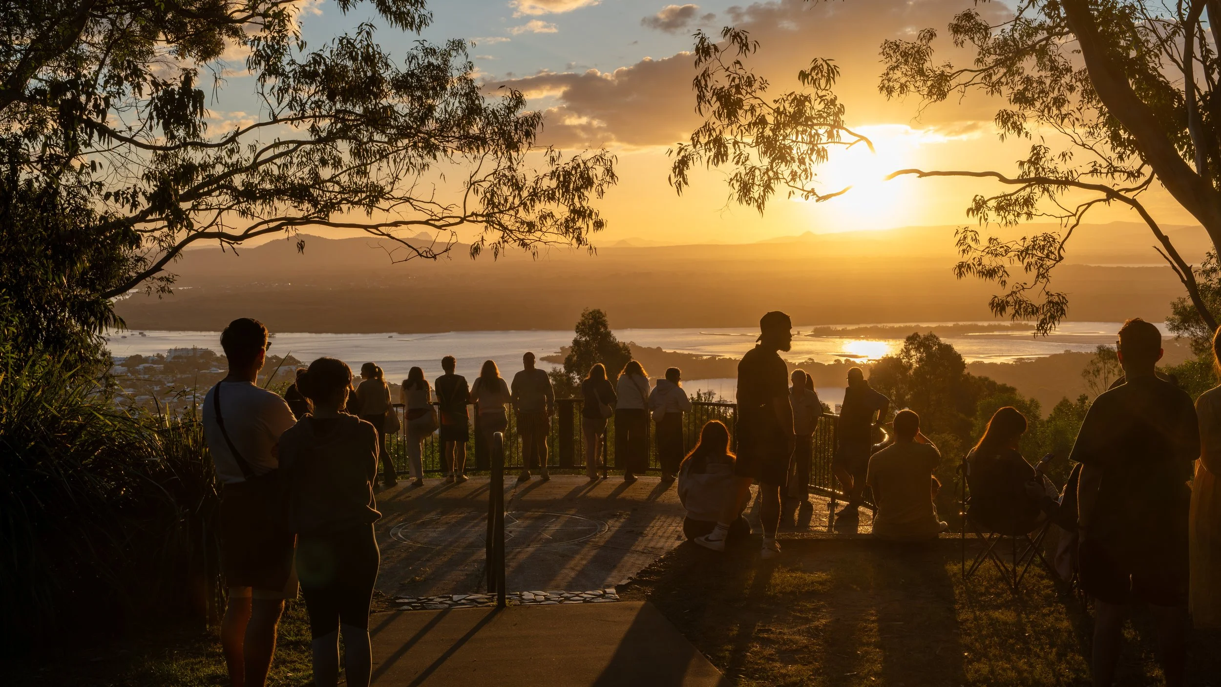 People watching the sunset from a scenic overlook with trees and a river in the background.