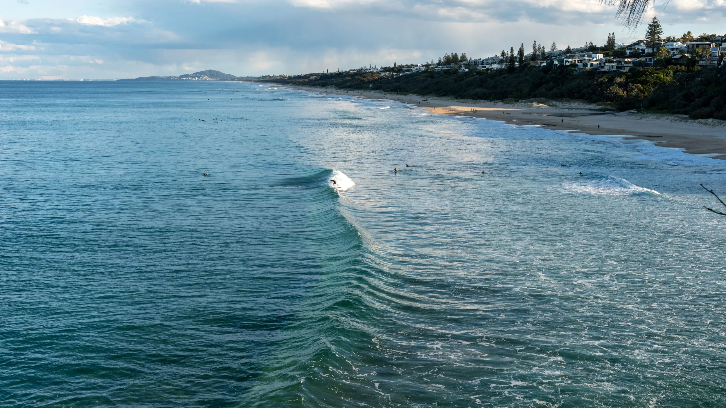 Ocean waves with surfers and a sandy beach in the background, residential homes on a hill, and cloudy sky.