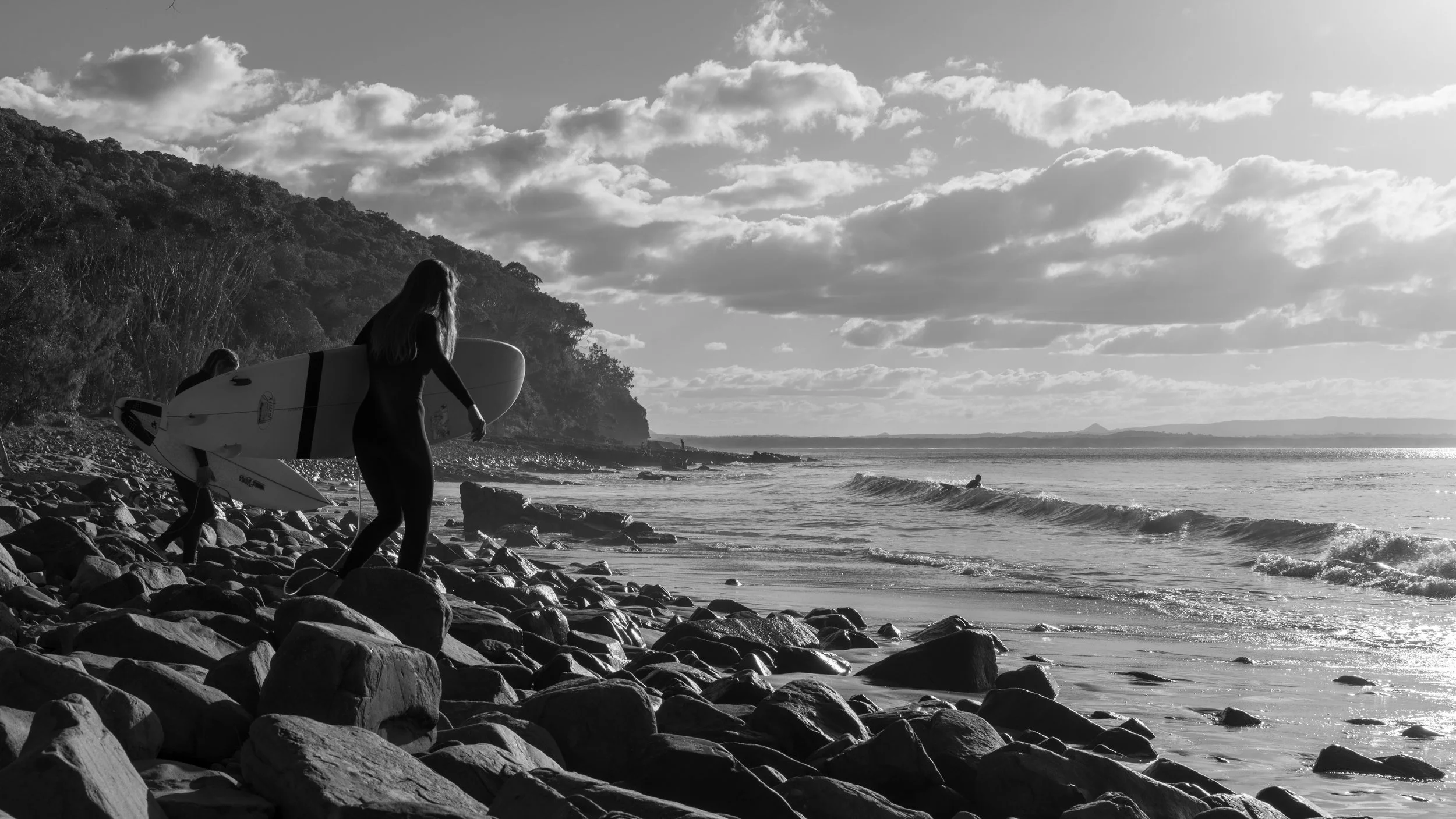 A person carrying a surfboard on a rocky beach near the ocean at sunset, with a cloudy sky and distant hills.