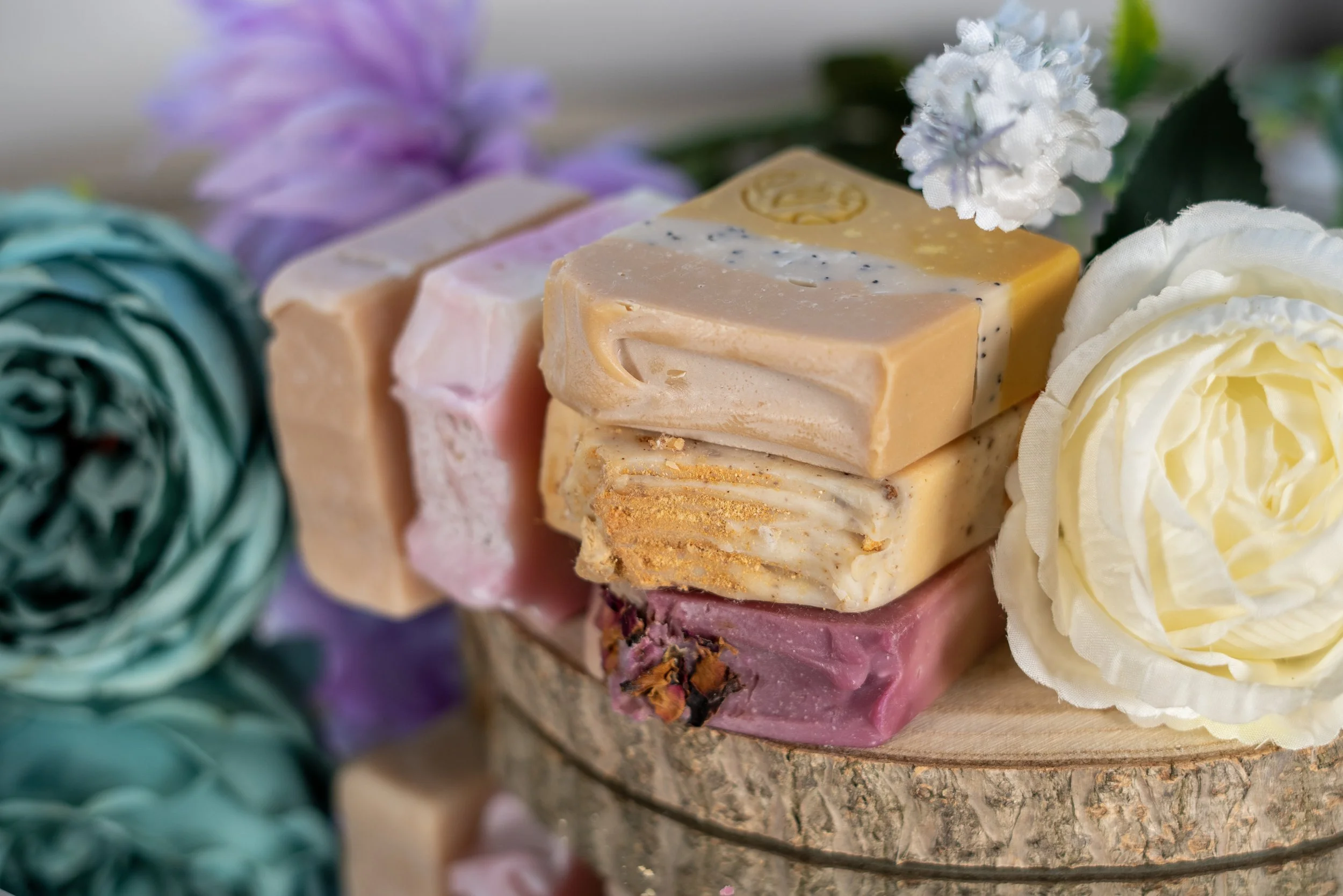 Stack of colorful handmade soap bars on a wooden slab with artificial flowers around, in pastel shades of pink, yellow, white, and beige.
