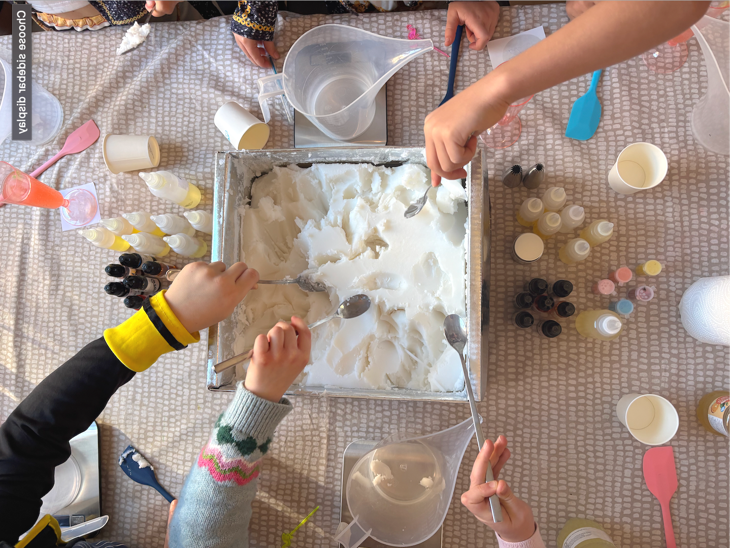 Children using spoons to scoop ice cream from a large tray during a dessert or ice cream party. Various colorful bottles, cups, and utensils are scattered across the table.