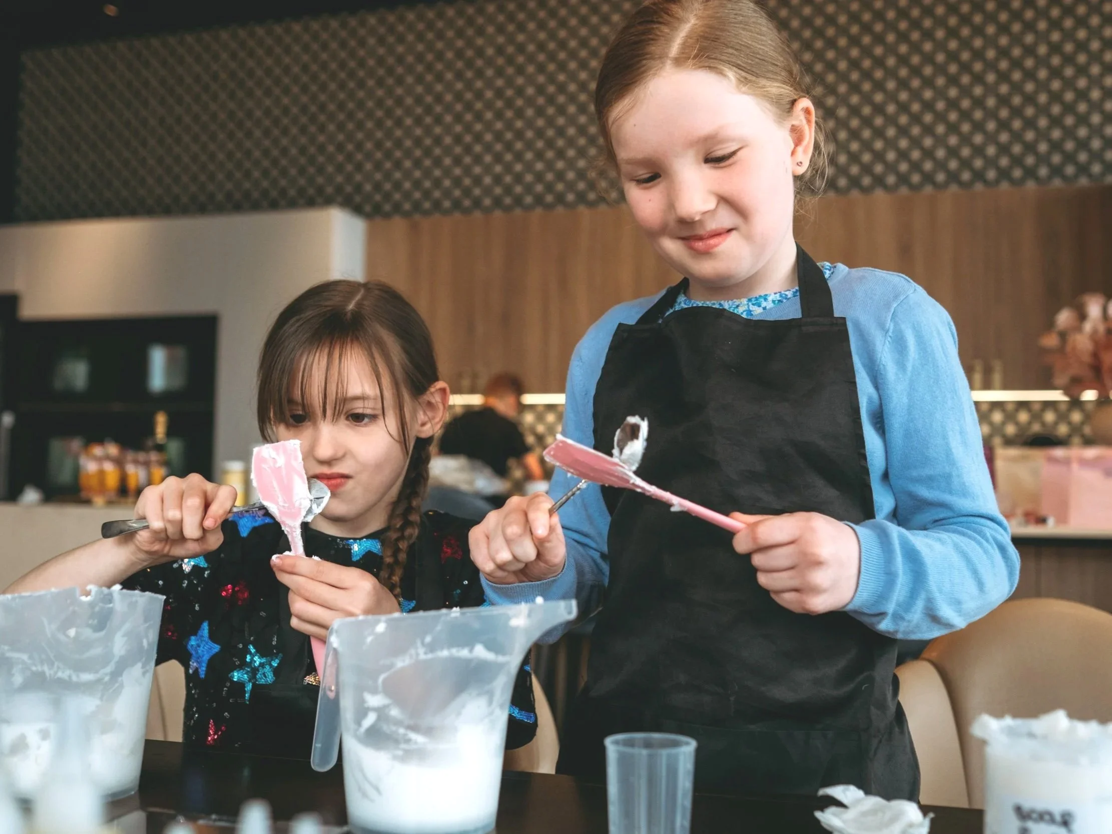 Two young girls are decorating cake with pink icing in a kitchen, one wearing a black apron and the other in a black shirt with colorful stars.