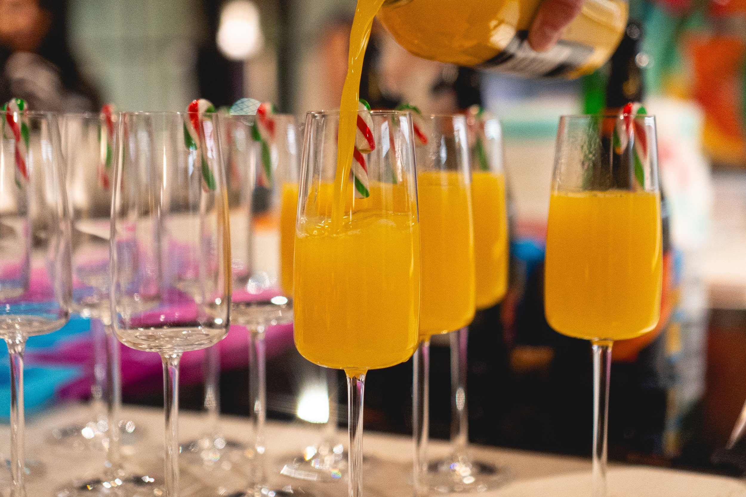 Glasses of orange juice being filled at a celebratory event with striped straws, on a decorated table.