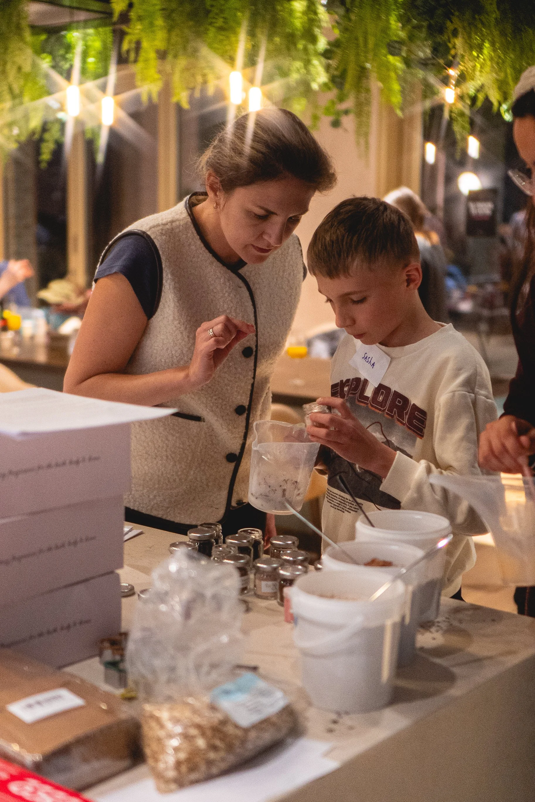 A woman and a boy look at small jars on a table at an indoor event. The woman has brown hair and wears a sleeveless beige vest, while the boy has short brown hair and a white sweatshirt. There are boxes, jars, and buckets on the table, with hanging lights and greenery above.