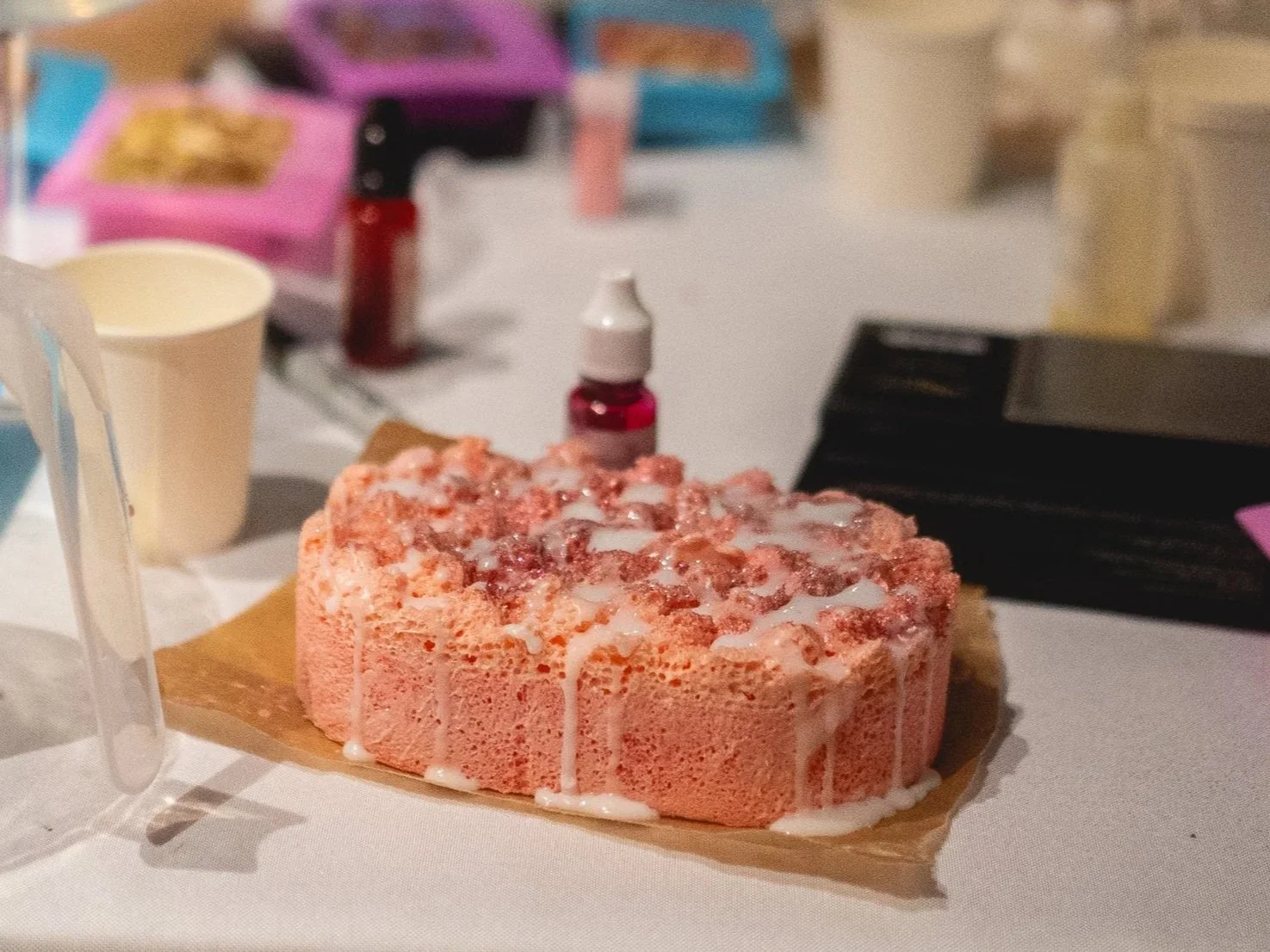 A pink soap-infused sponge placed on a table with various items in the background, including a cup, small bottles, and boxes.
