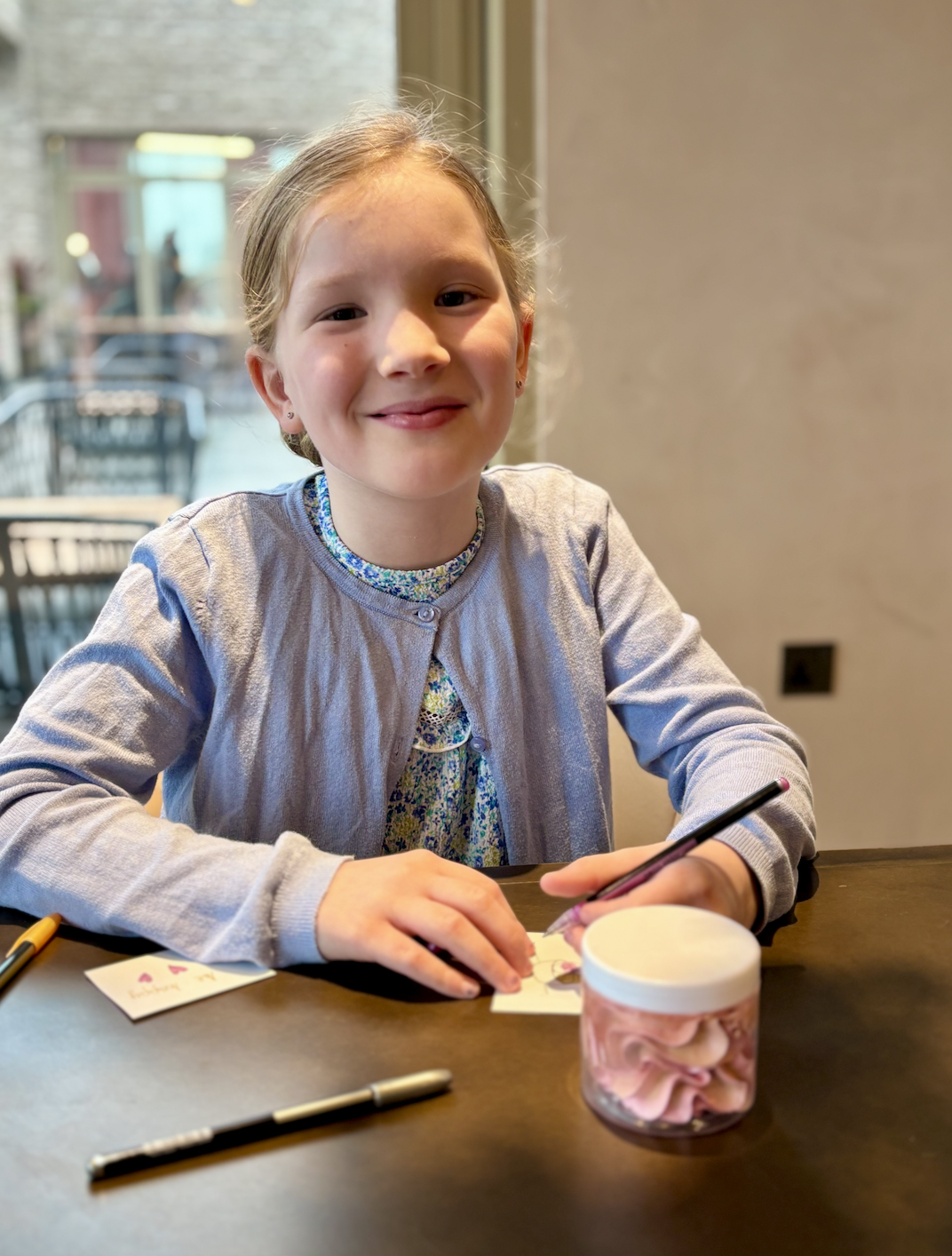 A young girl with blonde hair, smiling, sitting at a wooden table with drawing supplies and a jar of pink marshmallows, inside a room with a window showing an outdoor patio.