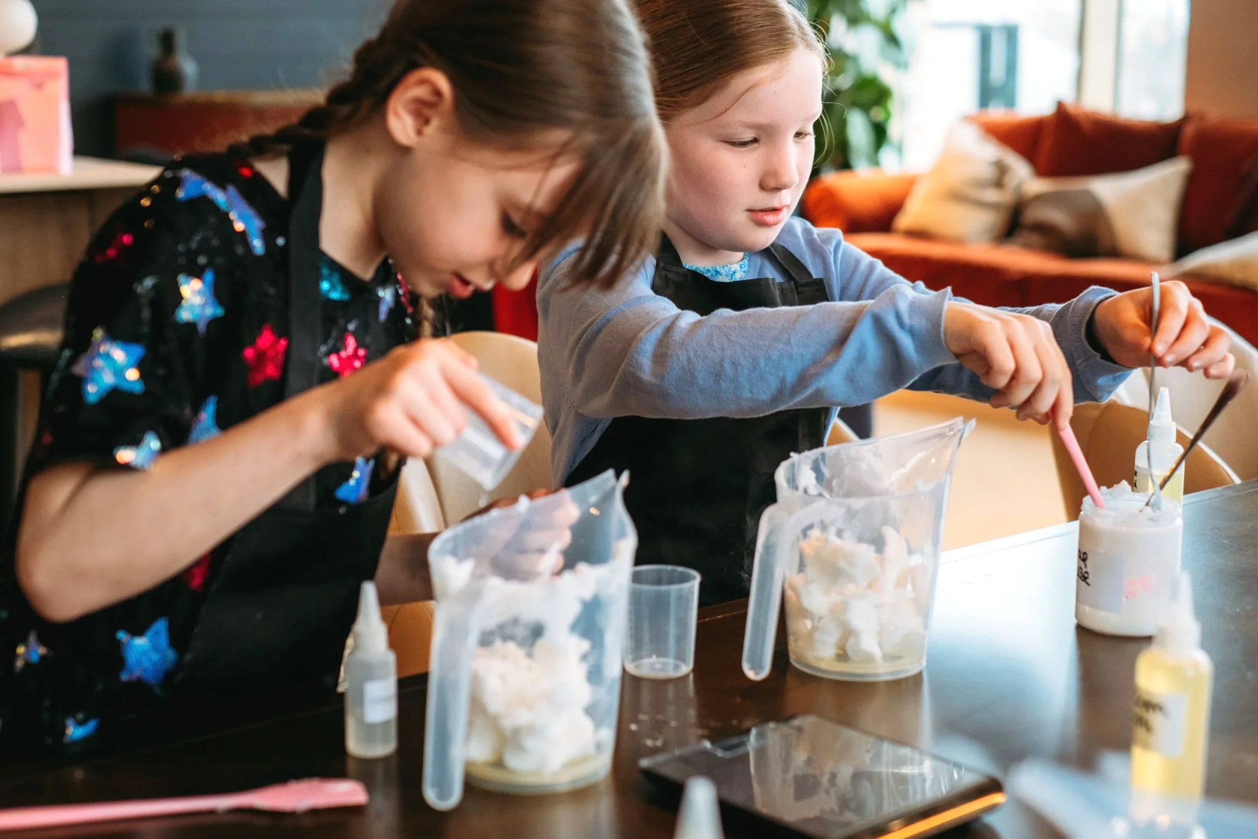 Two young girls creating slime at a table with containers, glue, and mixing tools in a cozy, well-lit room.