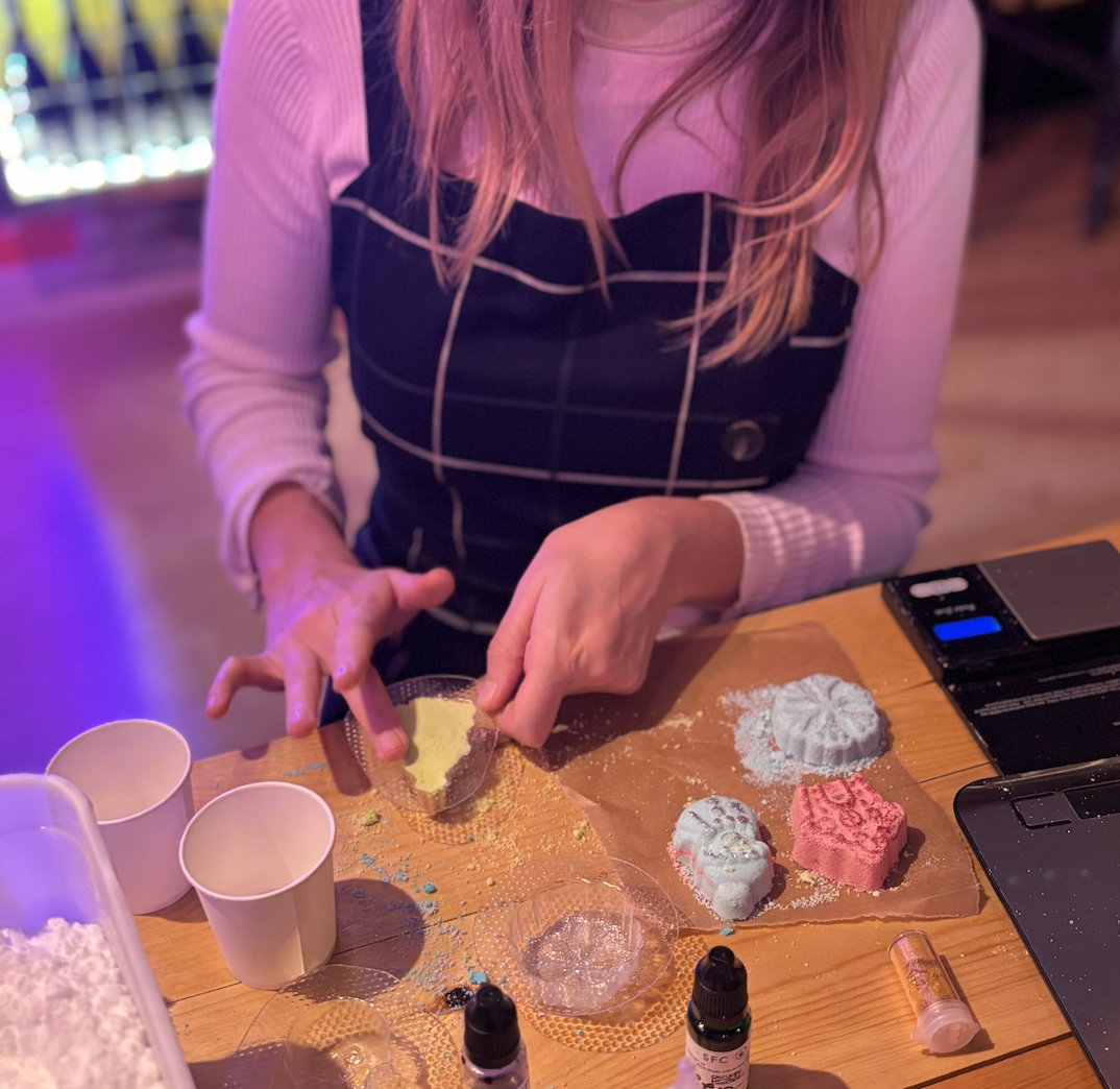 A person making bath bombs with colourful glitters and moulds on a wooden table, surrounded by small cups, oils, and tools.