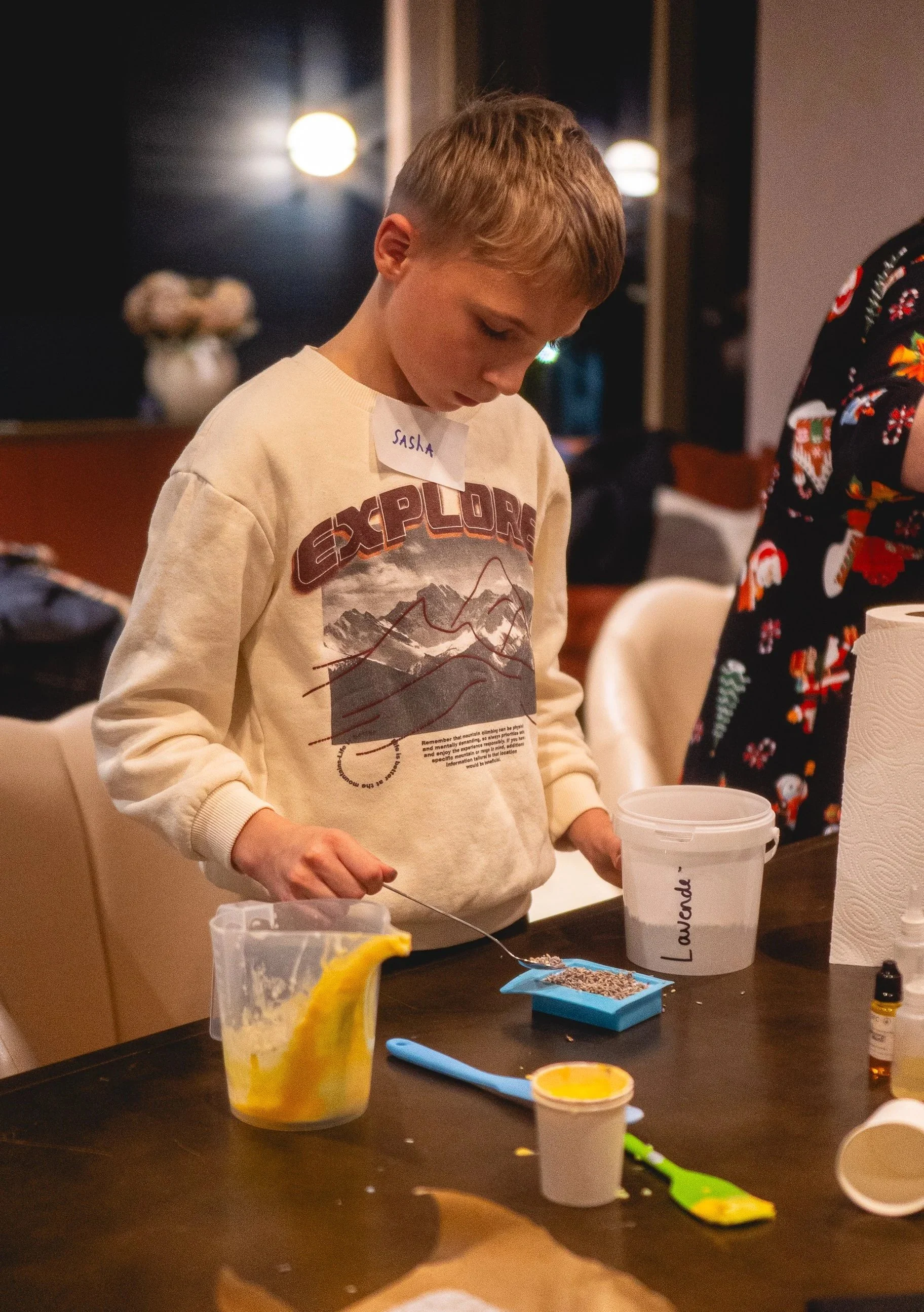 A young boy with a name tag reading 'Sasha' is pouring a substance from a spoon onto a blue surface on a table. The table has various items including a container labeled 'Lavender', a little yellow spatula, a small jar, and a bottle of essential oil.
