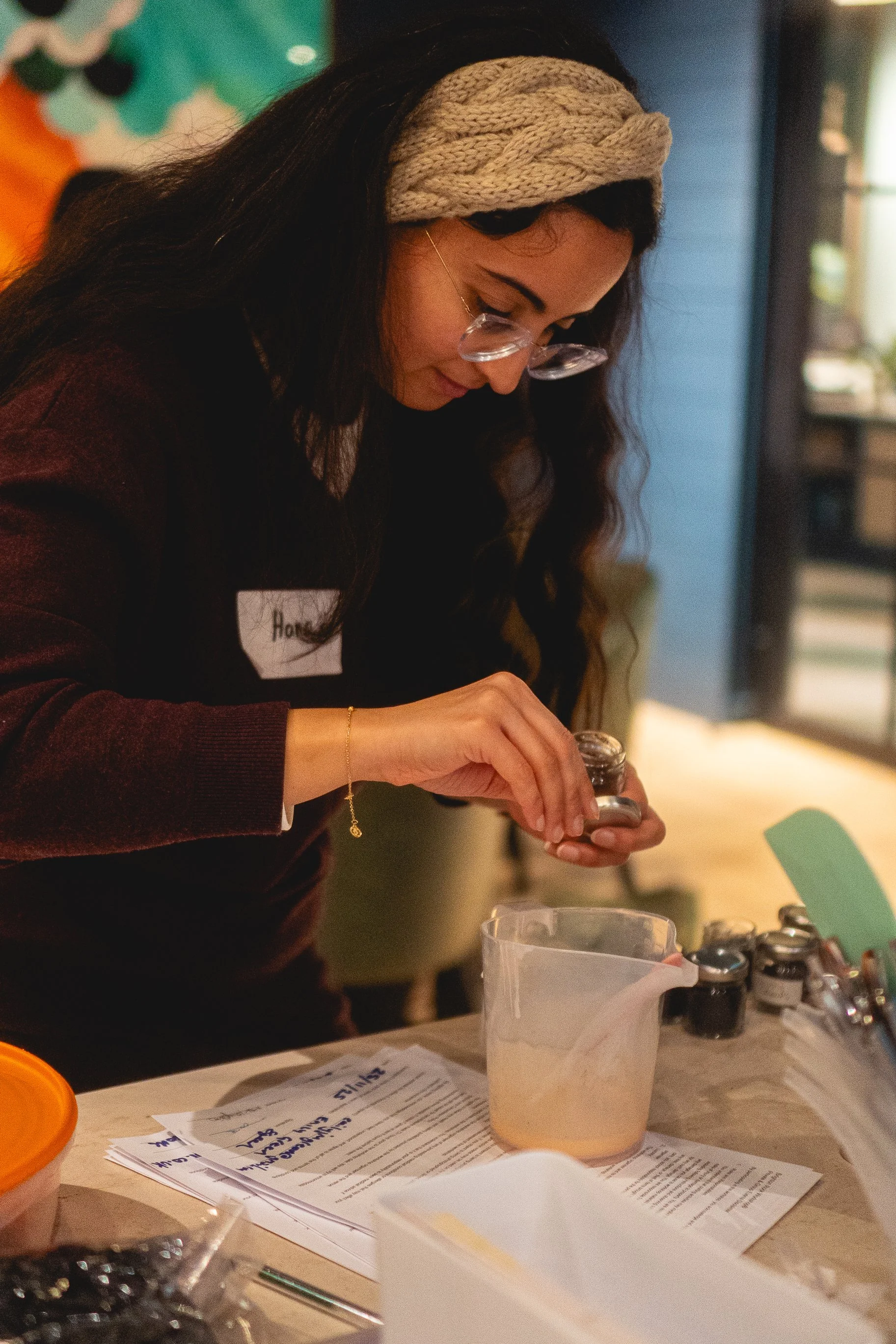Woman with long dark hair, glasses, and a beige knitted headband, working with small jars of paint or ink at a desk with papers and containers.