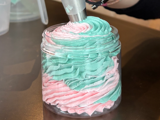 A glass jar filled with pastel pink and blue whipped cream, topped with a metallic object, on a wooden surface.