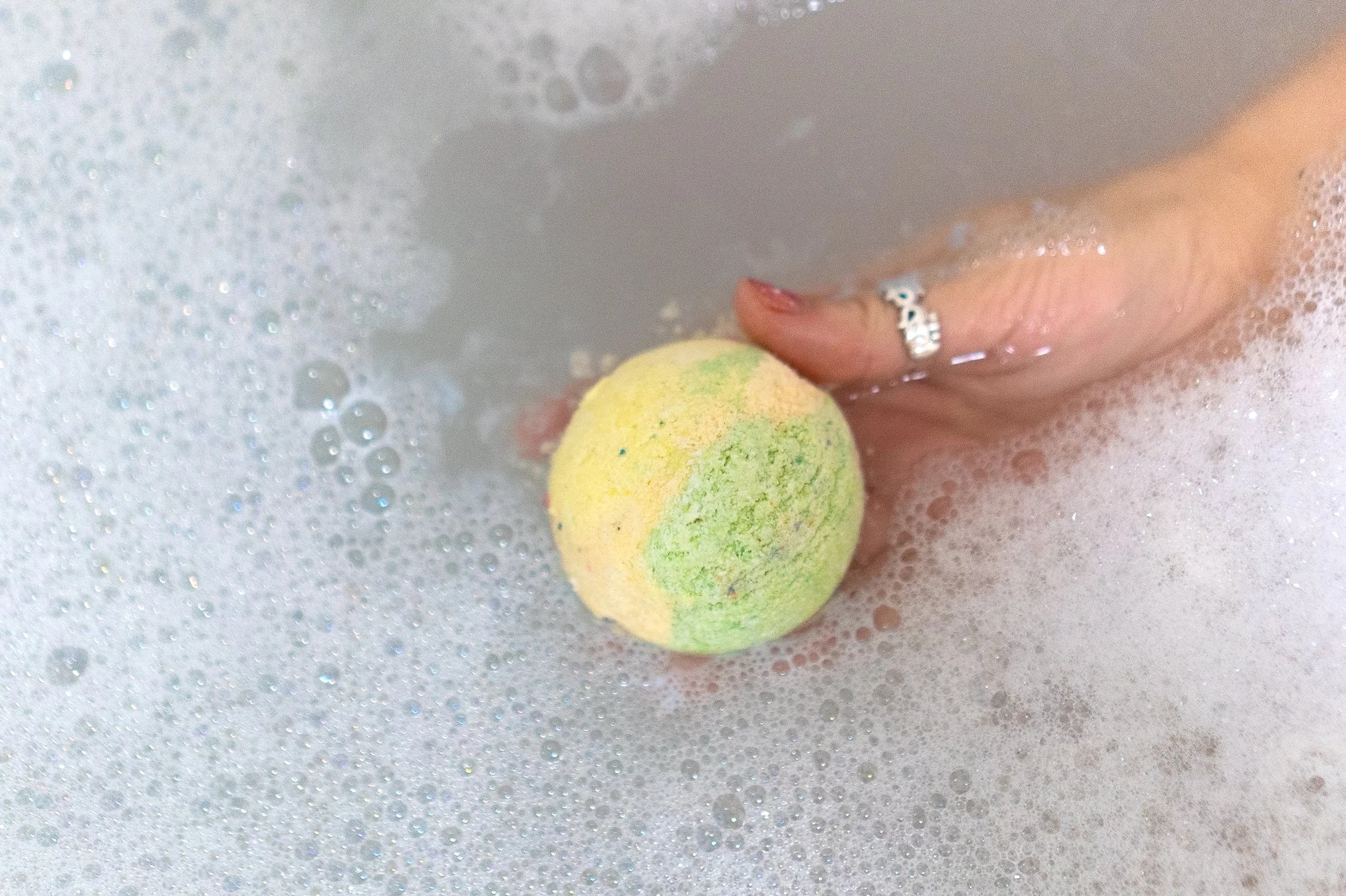 Person holding a pastel yellow and green bath bomb in a bubbly bathtub.