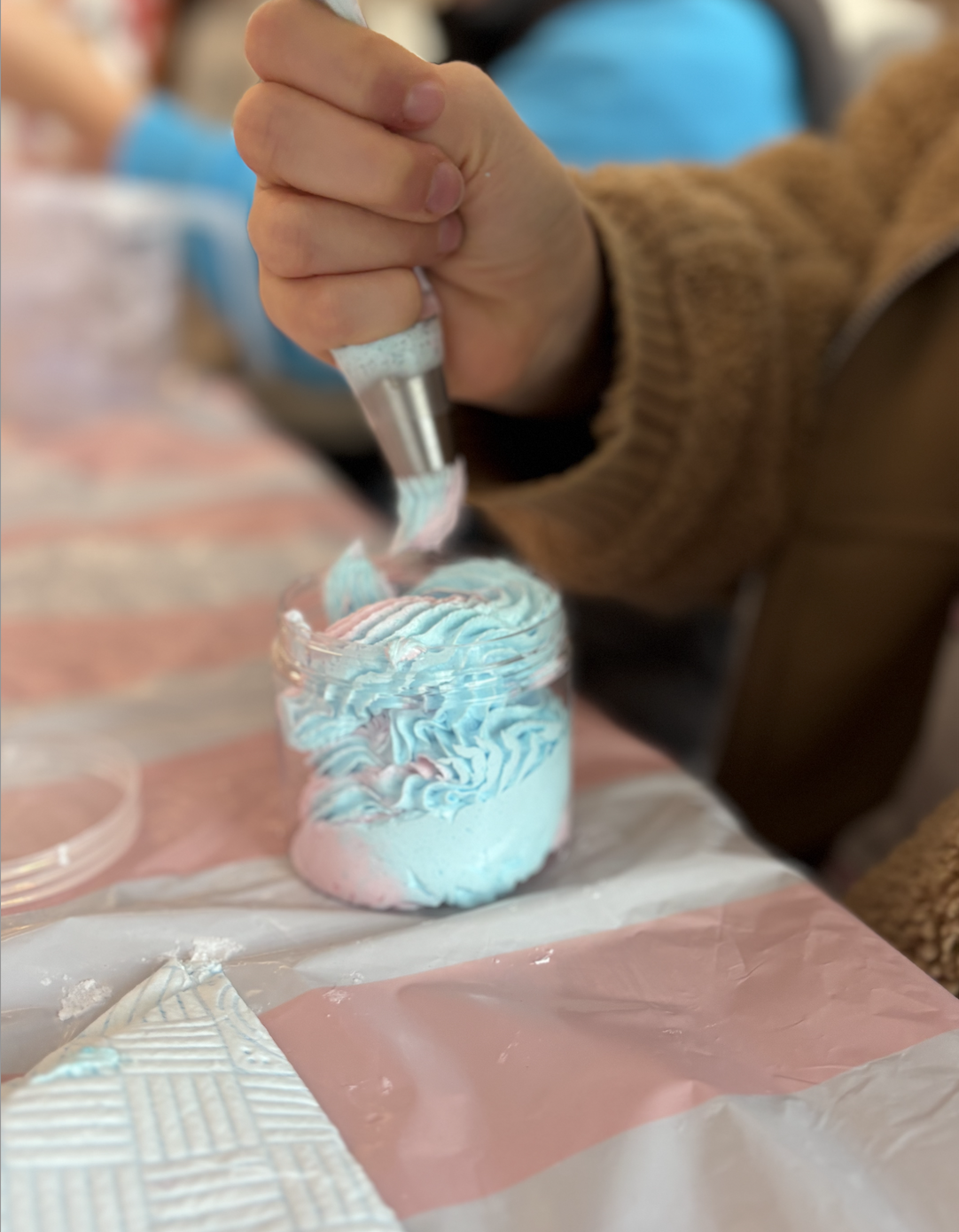 Close-up of a person's hand decorating a small jar with light blue and white icing with a piping nozzle, on a table covered with a pink and white tablecloth.