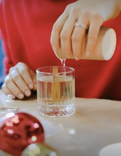 A woman pouring hot wax from a container into a glass candle vessel on a wooden surface, with Christmas ornaments nearby.