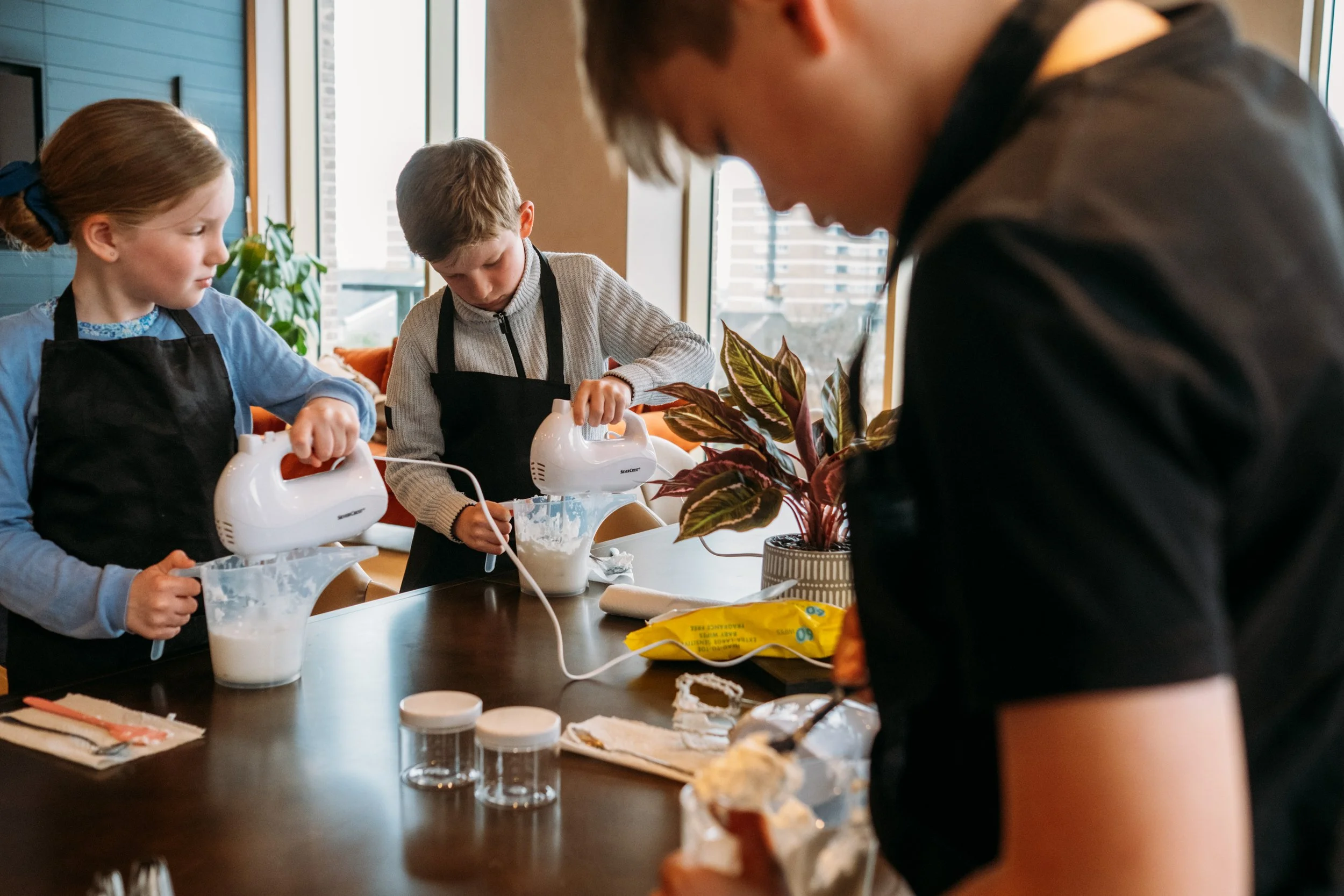 Three children and an adult in a kitchen, wearing aprons, using handheld electric mixers to prepare a baking mixture on a wooden table with baking ingredients and utensils.