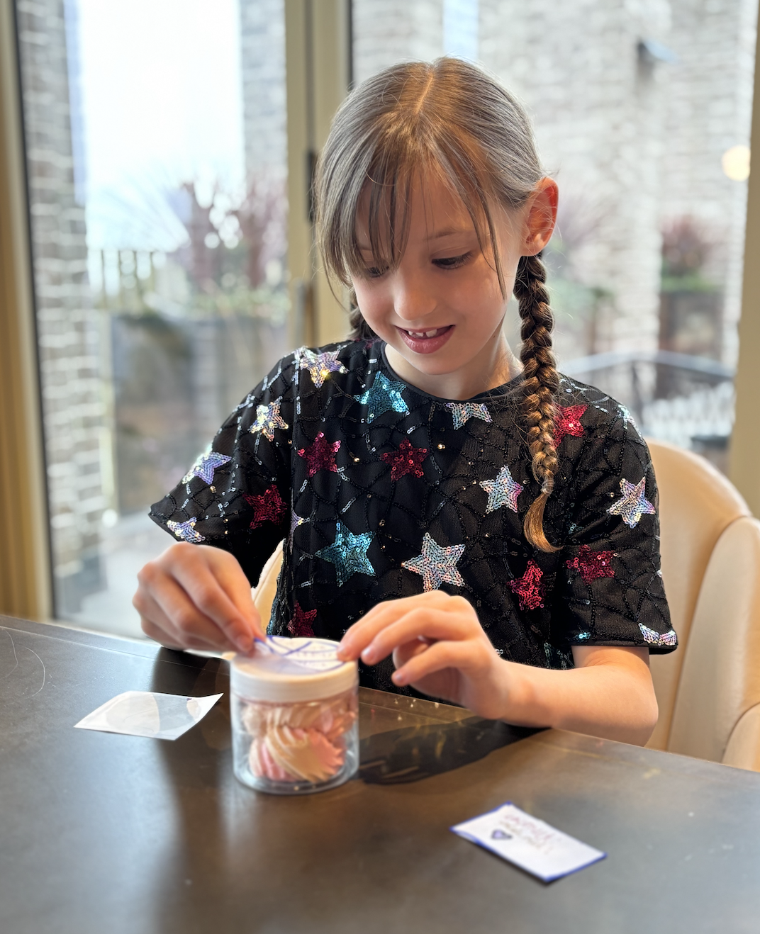 A young girl with braided hair, wearing a black dress with colorful star patterns, sitting at a table opening a small jar of cupcakes with pink and white swirls, in a bright room with large windows and outdoor view.