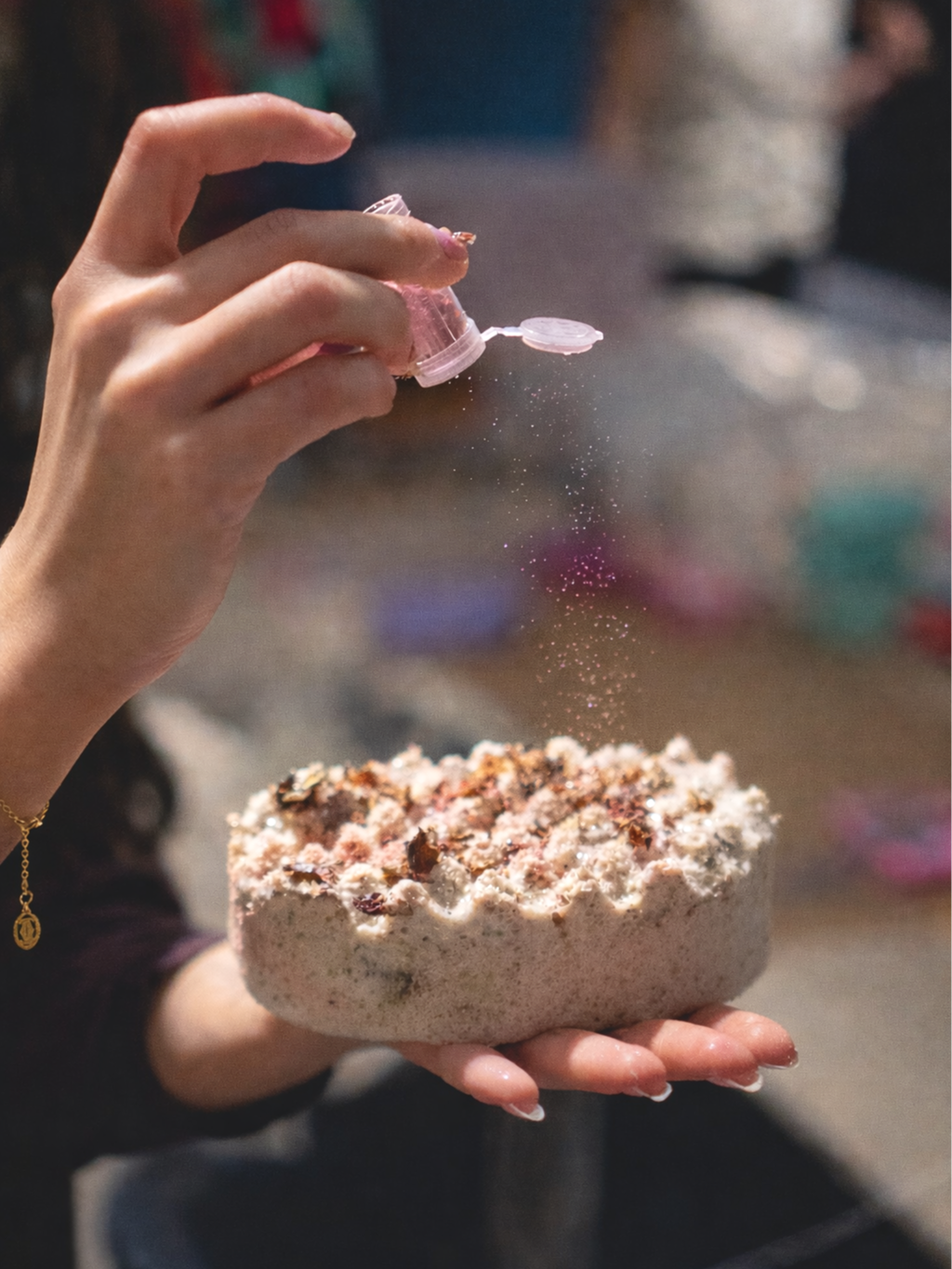 Person sprinkling pink glitter onto a small cake or dessert with a crackled surface, held in their hand.
