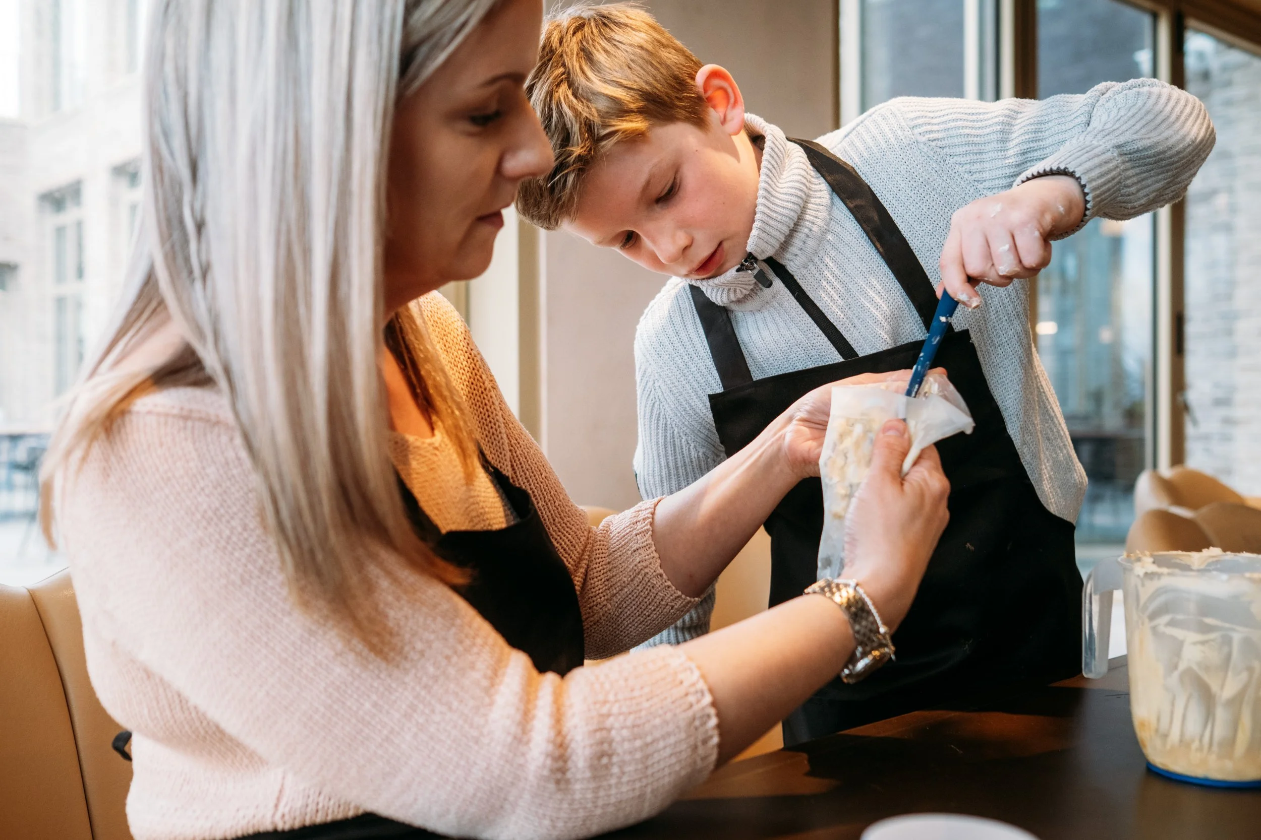 A woman and a young boy baking together in a kitchen, pouring batter into a bag with a piping tip.
