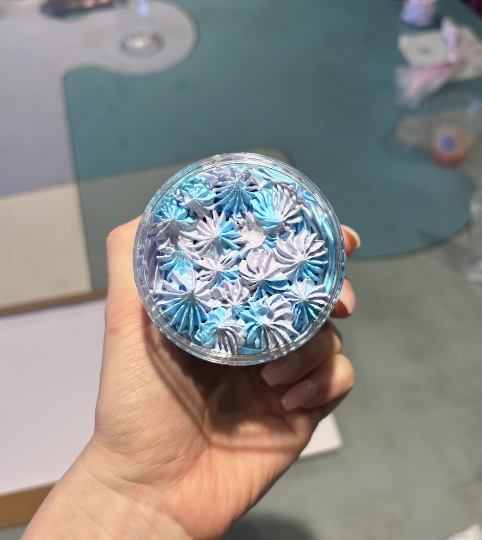 A hand holding a clear plastic cup filled with blue and purple swirled whipped cream or frosting, with a table and some paper or napkins in the background.