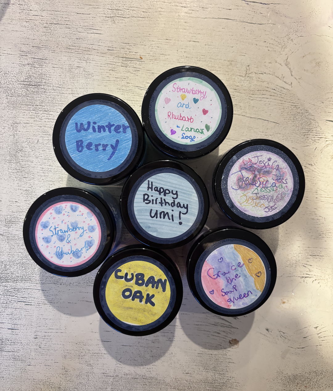 A collection of small jars with colorful handwritten labels on the lids, including flavors like Winter Berry, Strawberry and Raspberry, Cubain Oak, and Grace the Soap Queen, arranged on a white wooden surface.