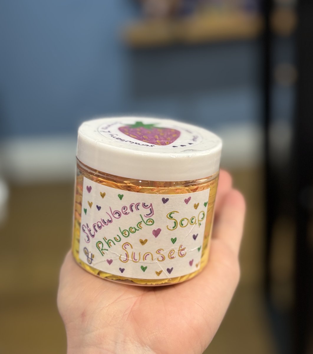 A hand holding a jar of strawberry and rhubarb soap with a white lid and a colorful handwritten label with small hearts.