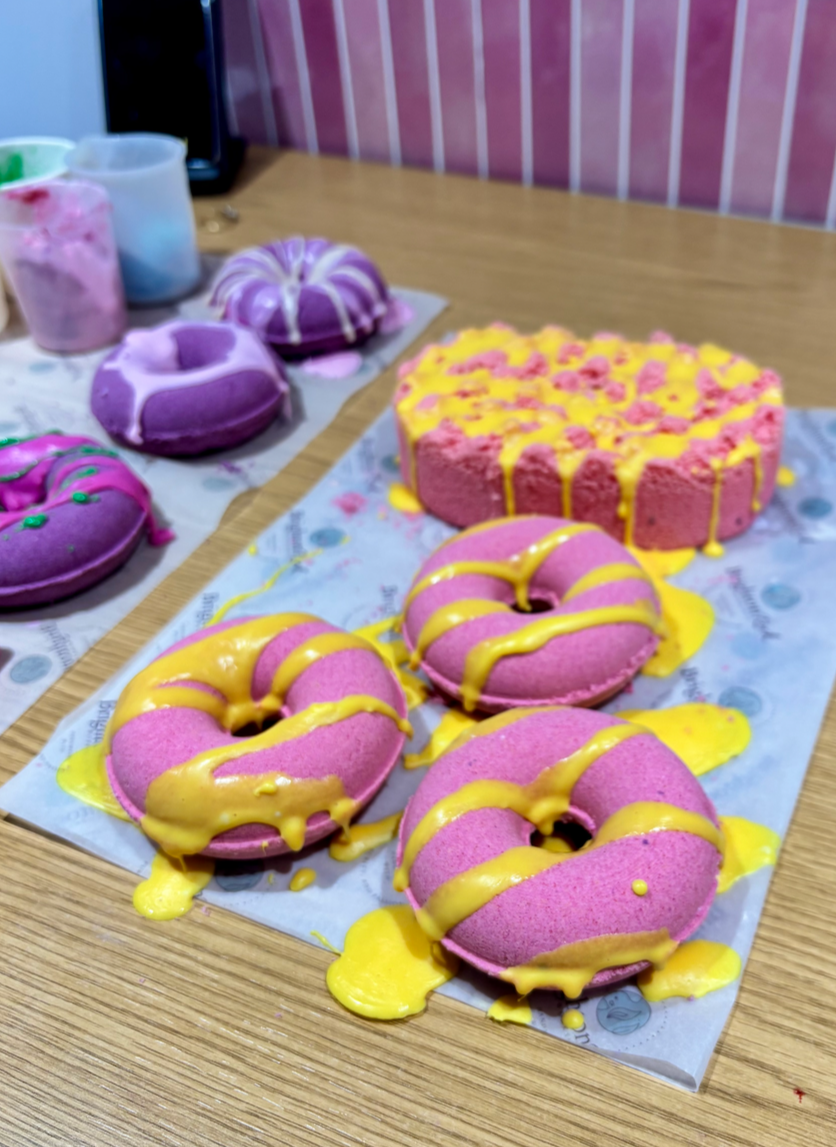 Pink donuts with yellow drizzle on display on a tray, along with a pink cake with yellow drizzle and purple macarons in the background.