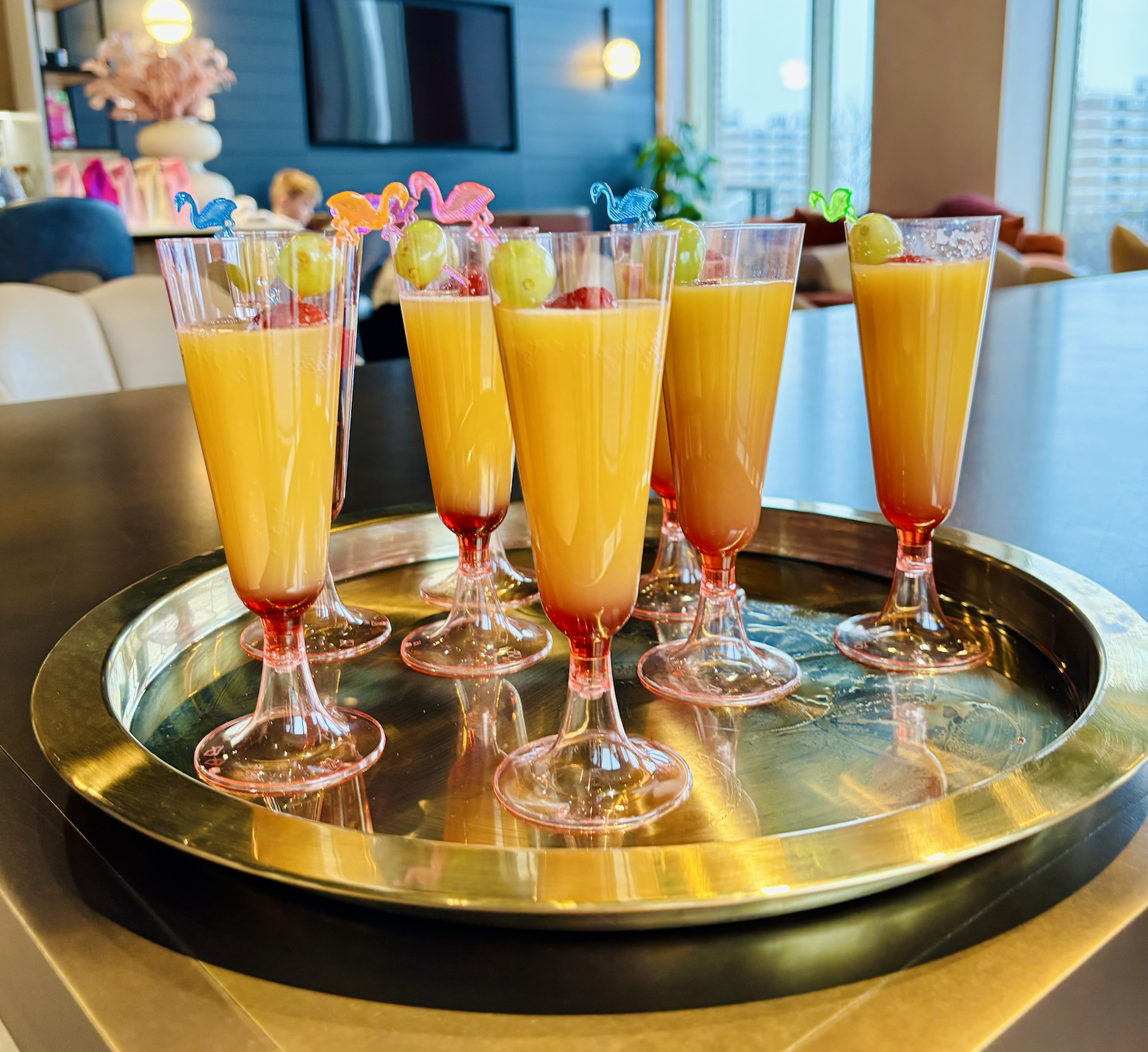 Six glasses of orange juice with grapes and colorful drink stirrers on a silver tray on a dining table in a bright, modern room.