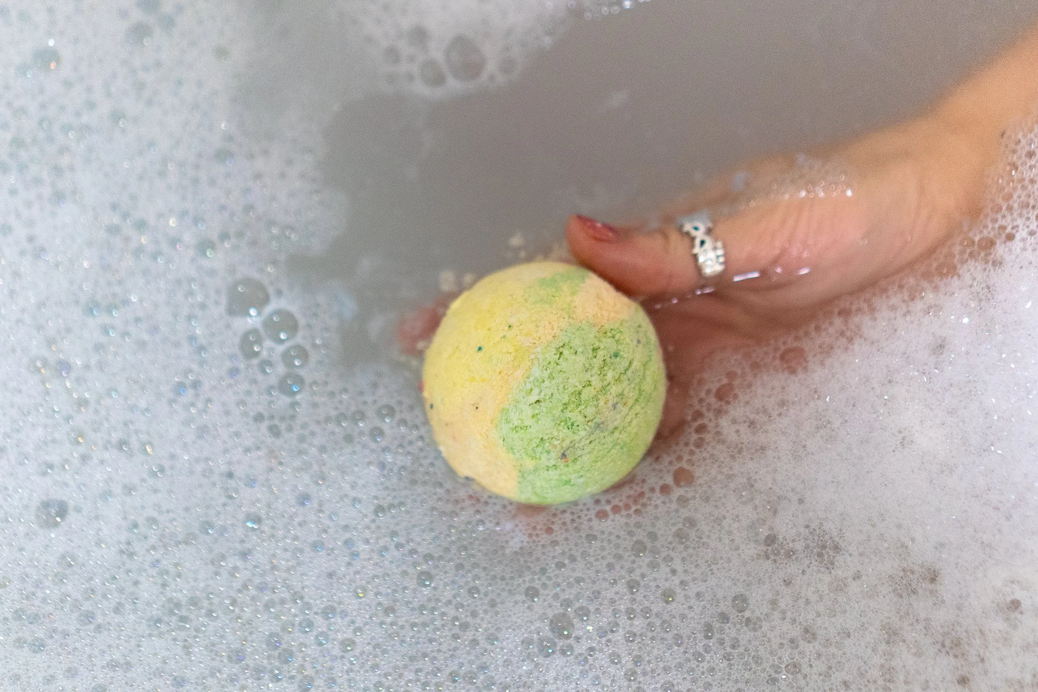 Person holding a yellow and green bath bomb in a bubbly bath.