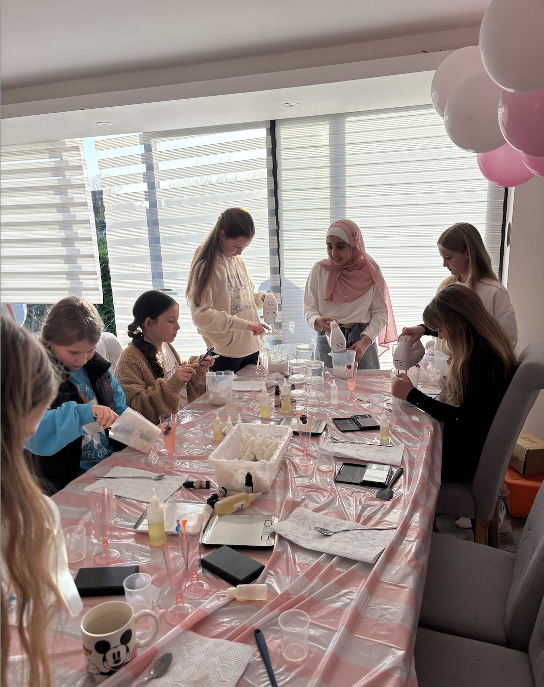 A group of young girls and women at a table engaged in a craft or baking activity with hand mixers, various supplies, and pink-themed decorations in a bright room.