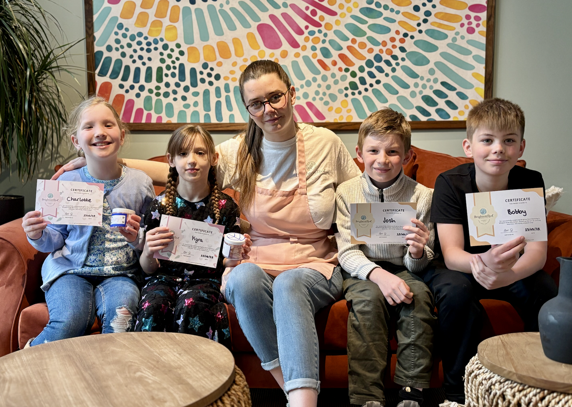 Group of four children and one adult woman sitting on a sofa in a room, holding certificates and small containers. The woman is wearing glasses and a pink apron. The children are smiling and sitting close together. There is a colorful abstract painti