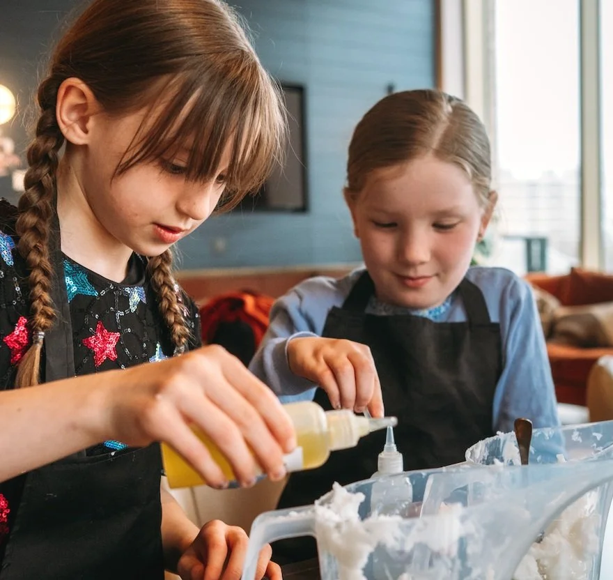 Two young girls wearing aprons are cooking or baking together at a table, using squeeze bottles to add ingredients to a container.