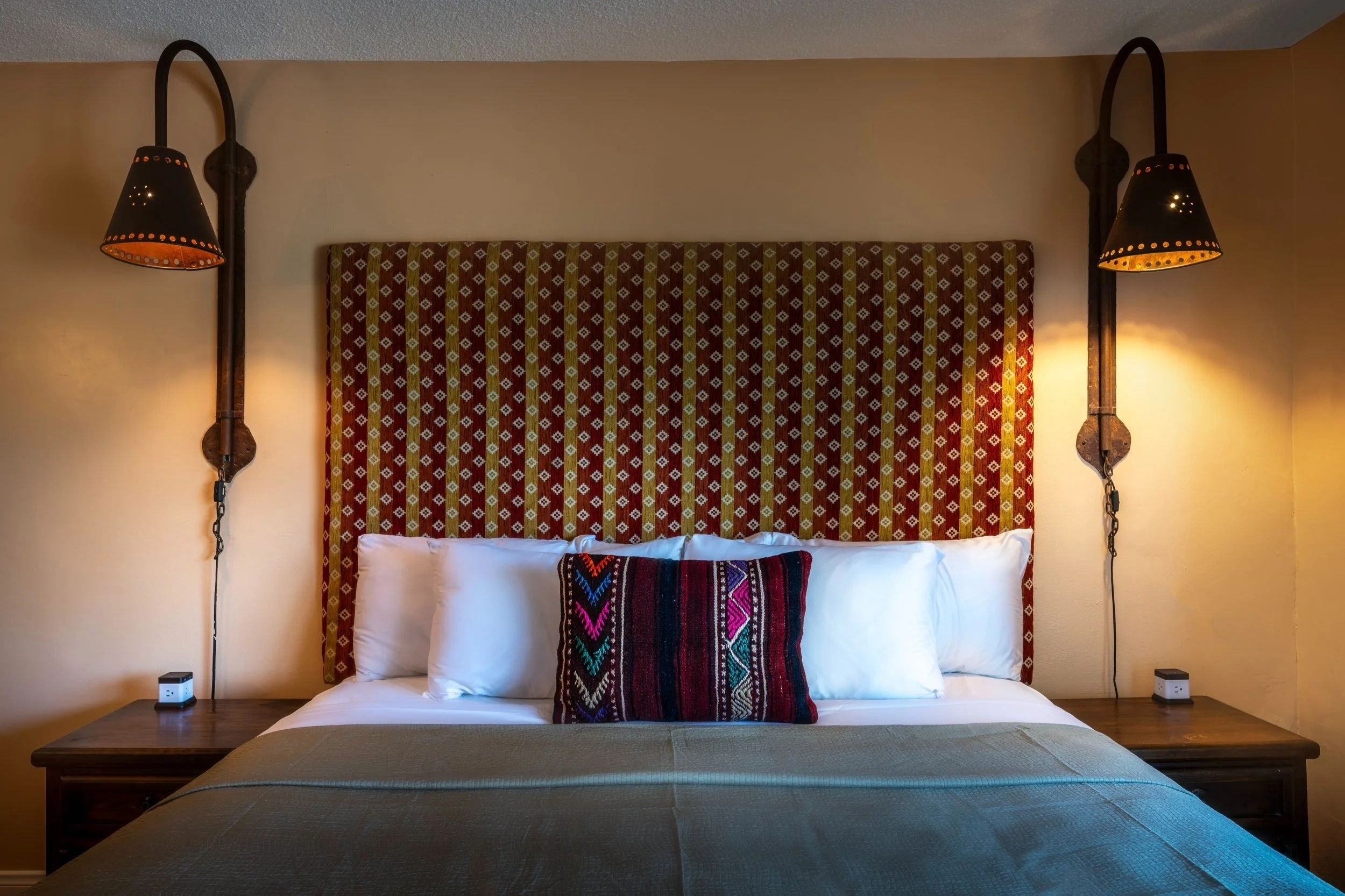 A bedroom with a bed that has a patterned fabric headboard, white pillows, a decorative pillow with colorful embroidery, and wooden side tables with wall-mounted black lamps on either side.