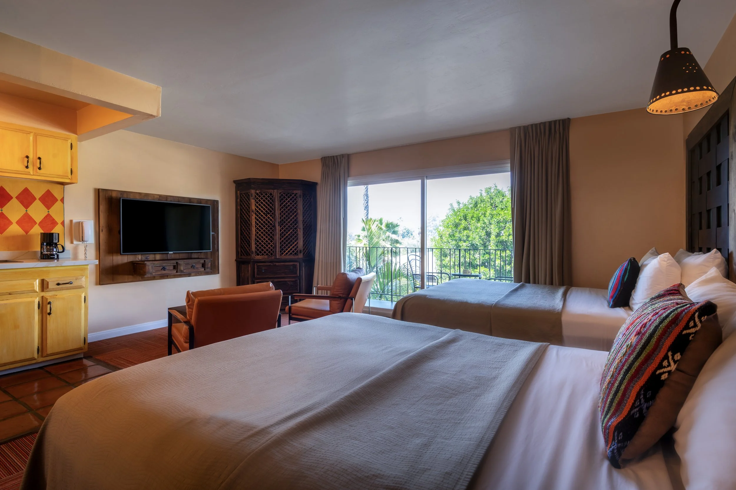 Hotel room with two beds, a balcony view of greenery, a television, two chairs, and a kitchenette area.
