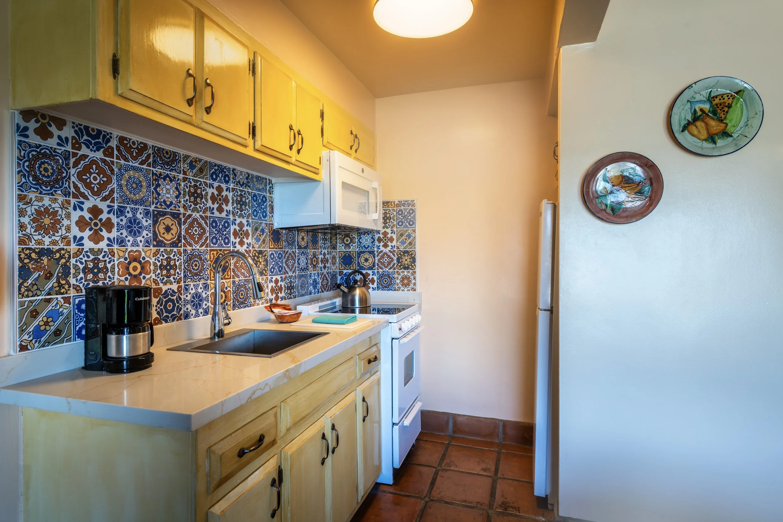 A small kitchen with yellow cabinets, a colorful patterned tile backsplash, a white microwave, a white stove, a coffee maker, a kettle, and decorative plates on the wall.
