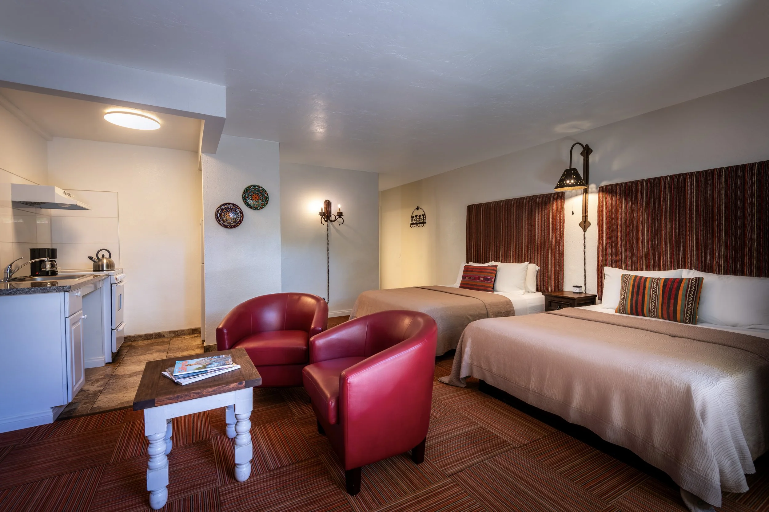 A hotel room with two double beds featuring striped headboards and pillows, a small wooden nightstand between the beds, red armchairs, a small coffee table with magazines, a kitchen, wall decorations, and warm lighting.
