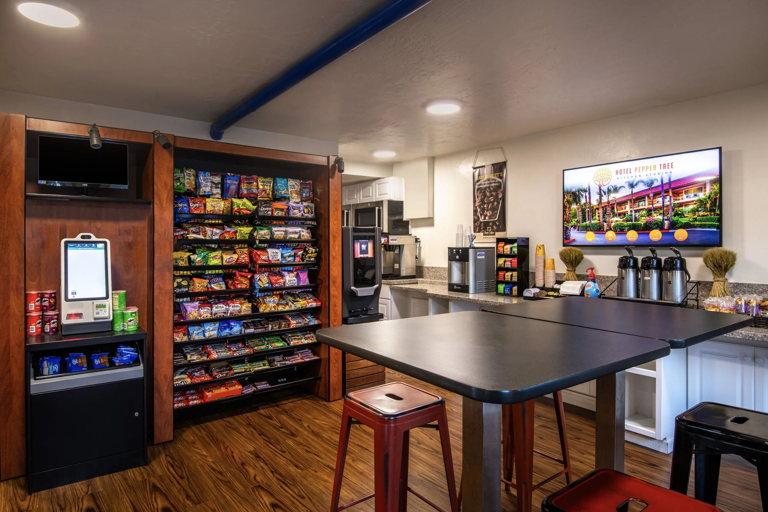 Snack station with a snack vending machine, coffee station, TV, tabletop, and food containers in a hotel breakfast room.